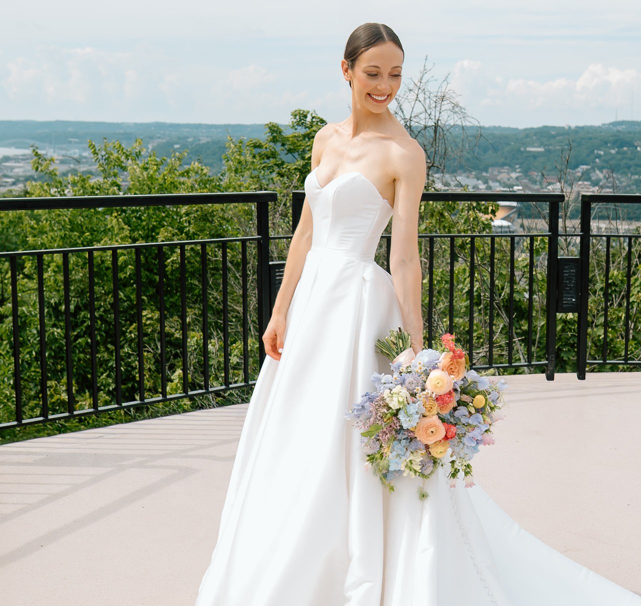 A woman in a white wedding dress holding a colorful bouquet, standing outdoors on a balcony with green trees and a cityscape in the background.