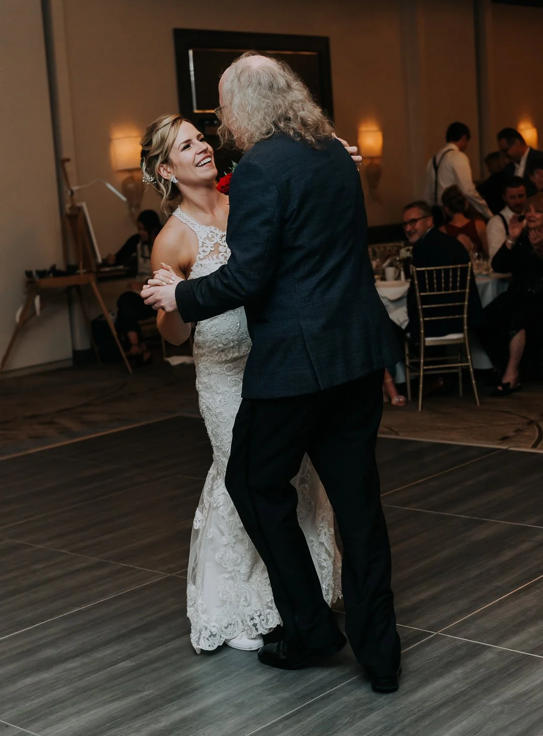 A bride and an older man dancing together at a wedding reception, smiling and holding hands, with guests seated at tables in the background.