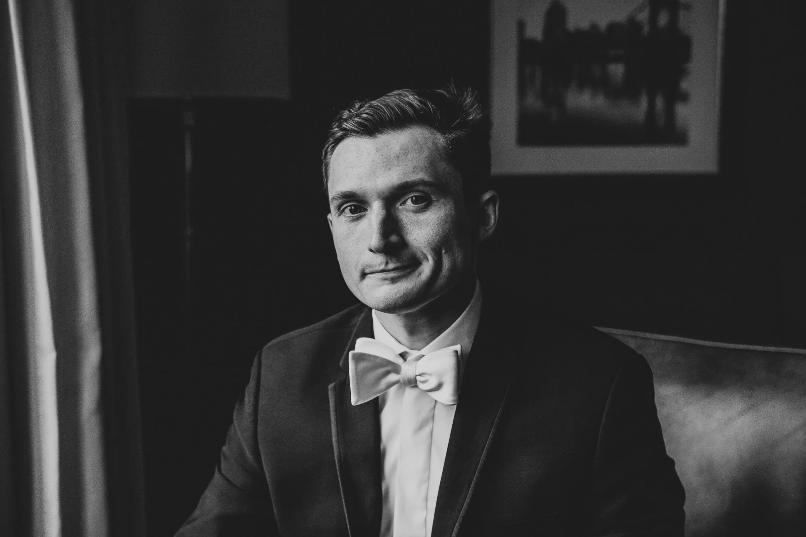 Black and white photo of a young man in formal attire with a bow tie, sitting indoors.