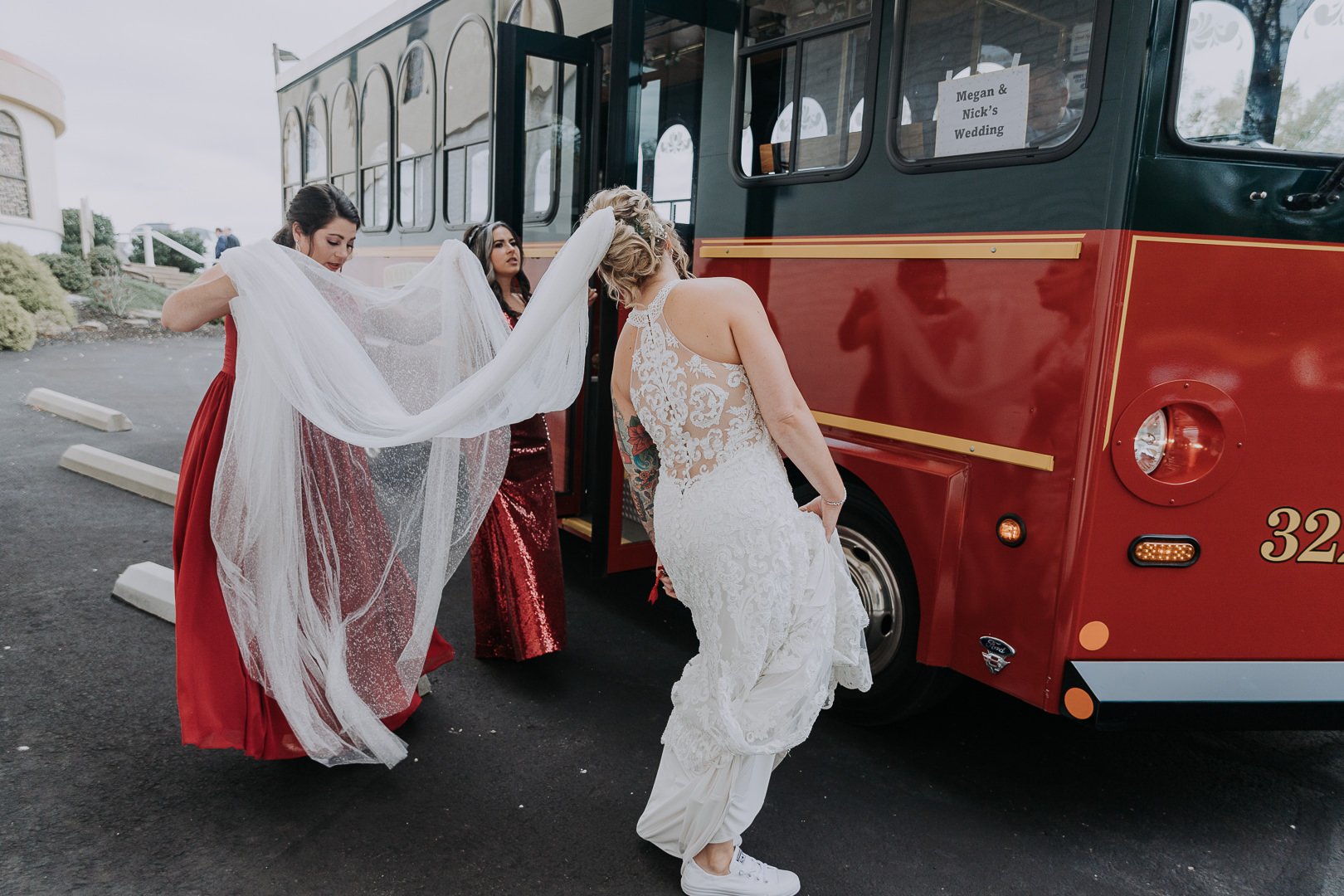 A bride in a white wedding dress and two bridesmaids in red dresses help the bride into a white pair of sneakers as she prepares to leave the bus for her wedding.