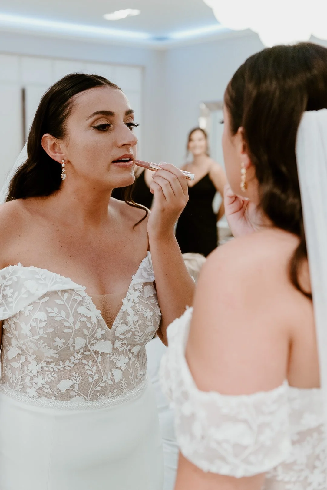 Bridal makeup artist applying lipstick to a bride while she looks in a mirror, with another woman in the background.