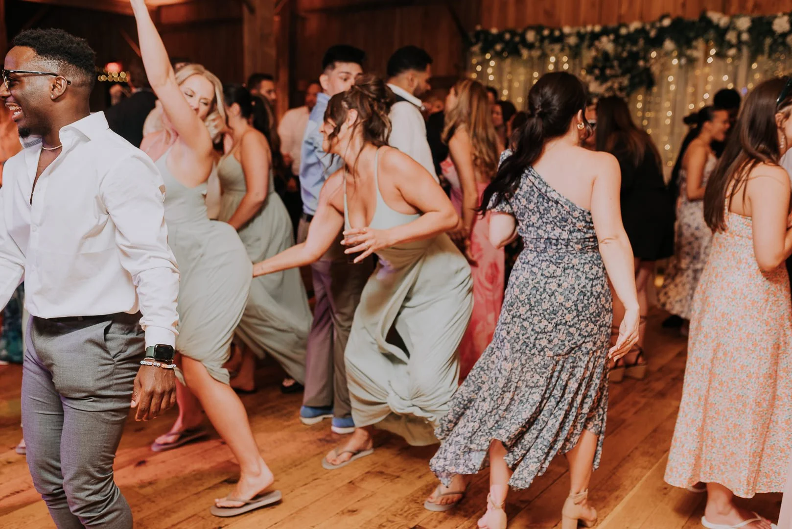 People dancing at a celebration party in a rustic hall decorated with string lights and floral arrangements.