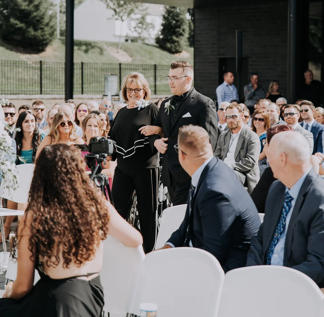 A young man in a black suit walking down an outdoor aisle, escorted by an older woman in black attire, at a wedding ceremony with seated guests in the background.