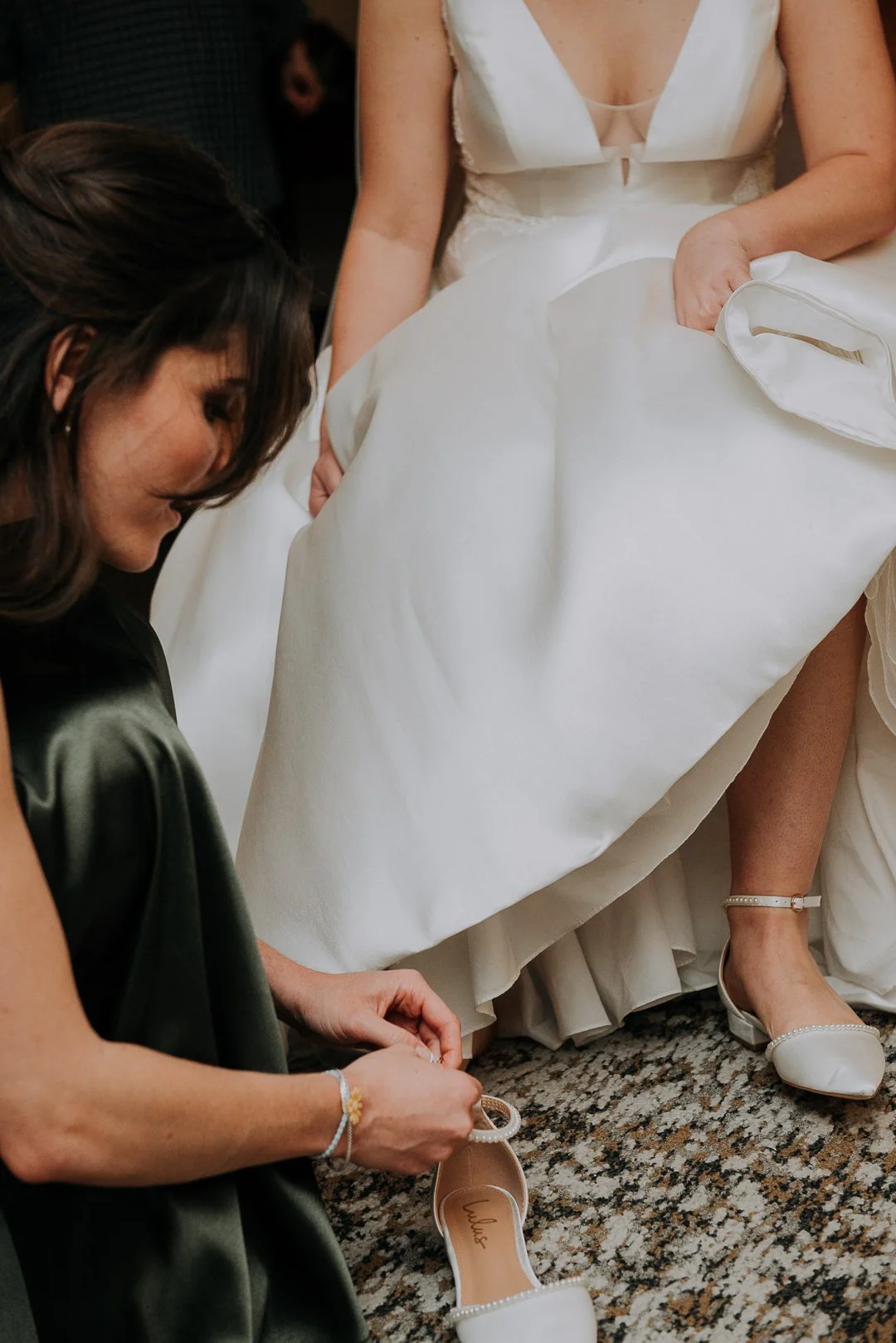 A bride in a white wedding dress sitting while another woman in a dark green dress helps her put on a white shoe.