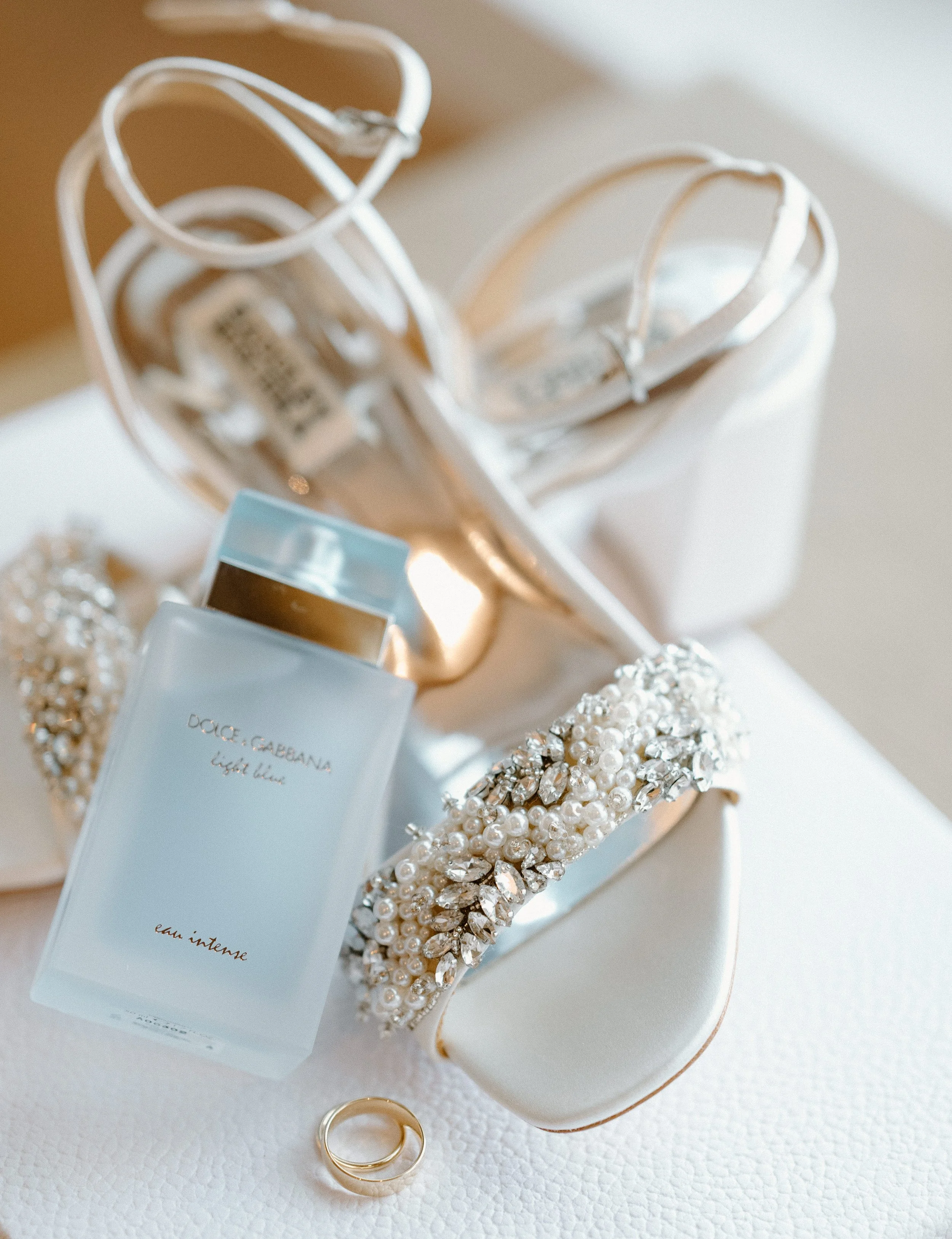 A white jewelry box with a pearl and rhinestone bracelet, a bottle of Dolce & Gabbana perfume, a gold ring, and a pair of white elegant high-heeled shoes with ankle straps decorated with rhinestones on a textured white surface.