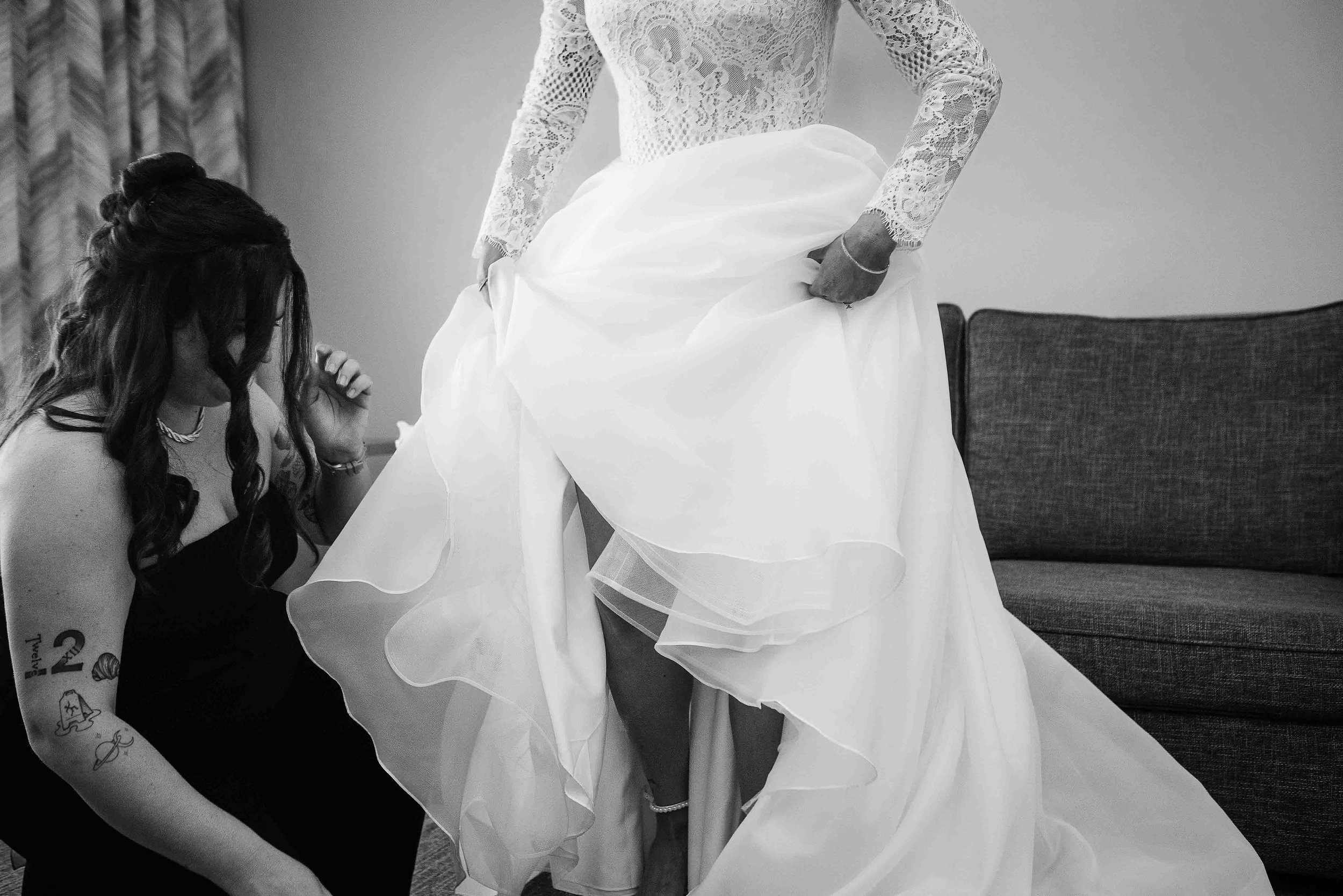 A woman wearing a bride's dress with lace long sleeves and a tulle skirt is having her dress arranged or adjusted. Another woman, with long dark hair and tattoos on her arm, is kneeling and assisting with the dress. The scene takes place indoors with