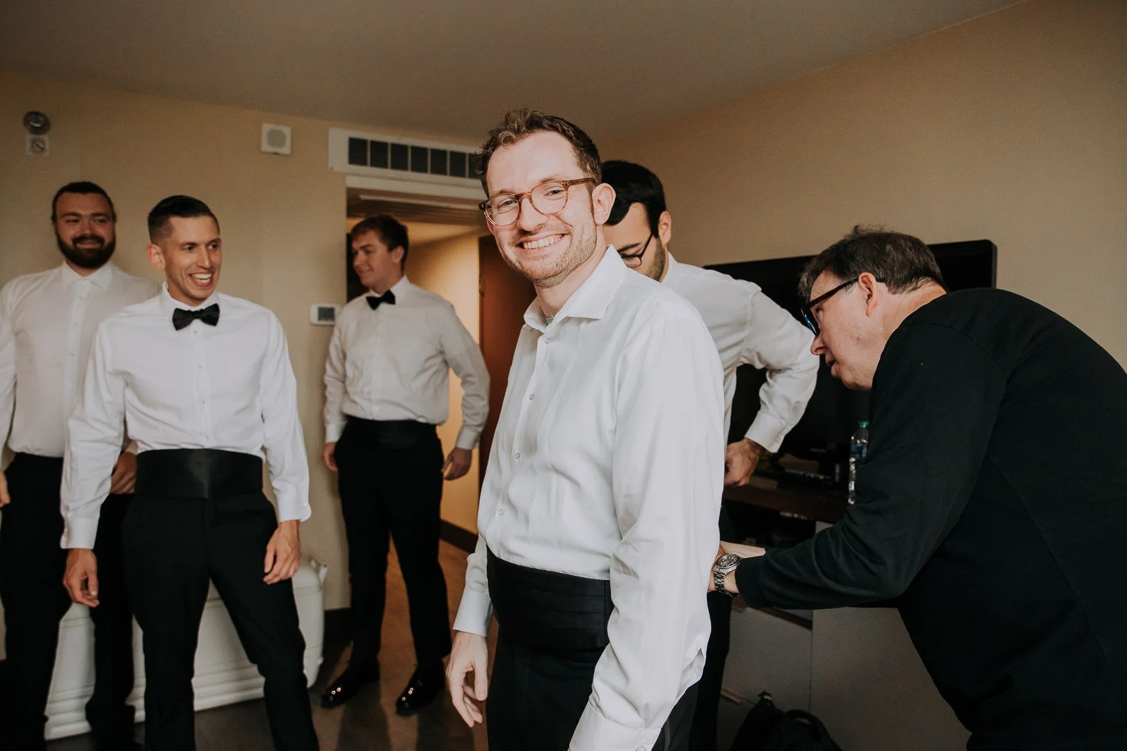 Group of men dressed in formal white shirts and black pants, preparing for a special occasion in a hotel room. One man in the foreground is smiling and looking at the camera, while others are adjusting their attire or talking in the background.