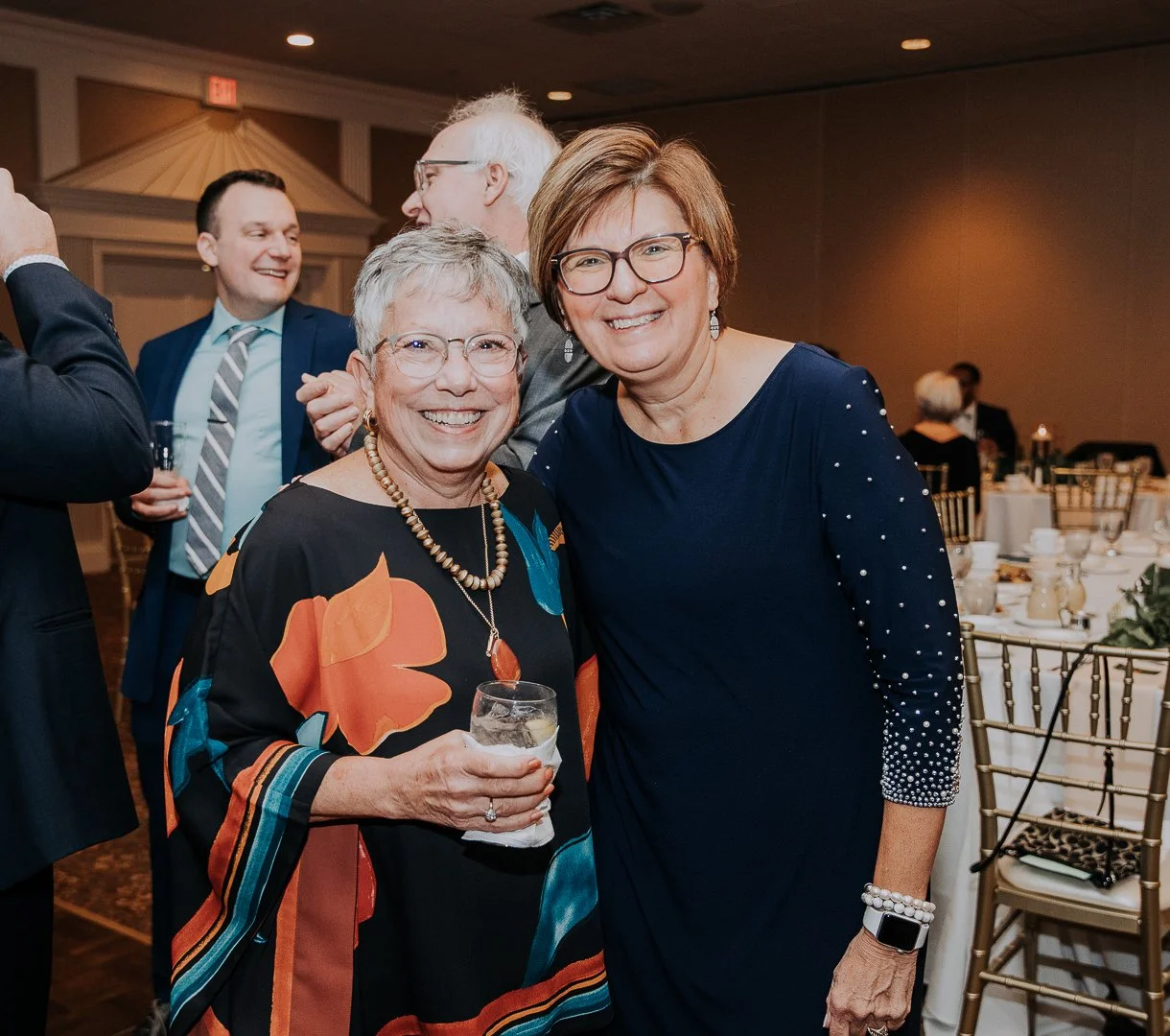 Two women smiling at a social event, one holding a drink, with tables and other guests in the background.