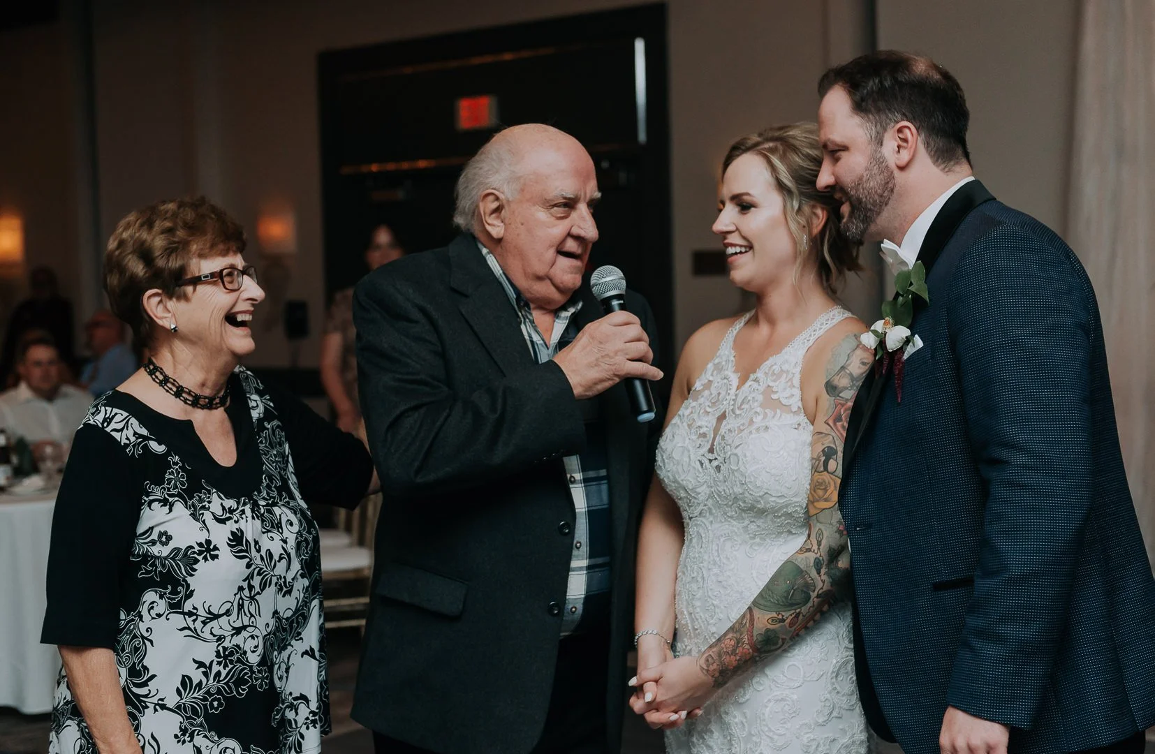 A wedding reception scene with a bride and groom holding hands, an older man speaking into a microphone, and a smiling woman in black and white attire; indoor setting with guests in the background.