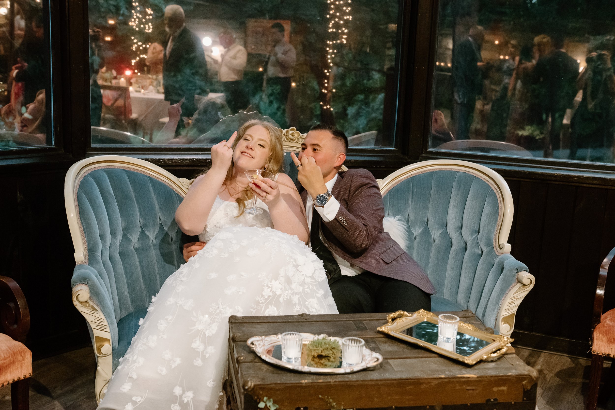A newlywed couple sitting on a vintage sofa at their wedding reception, with the bride holding a drink and making a playful gesture, and the groom also making a gesture near his face. Guests are visible through the window behind them, celebrating and