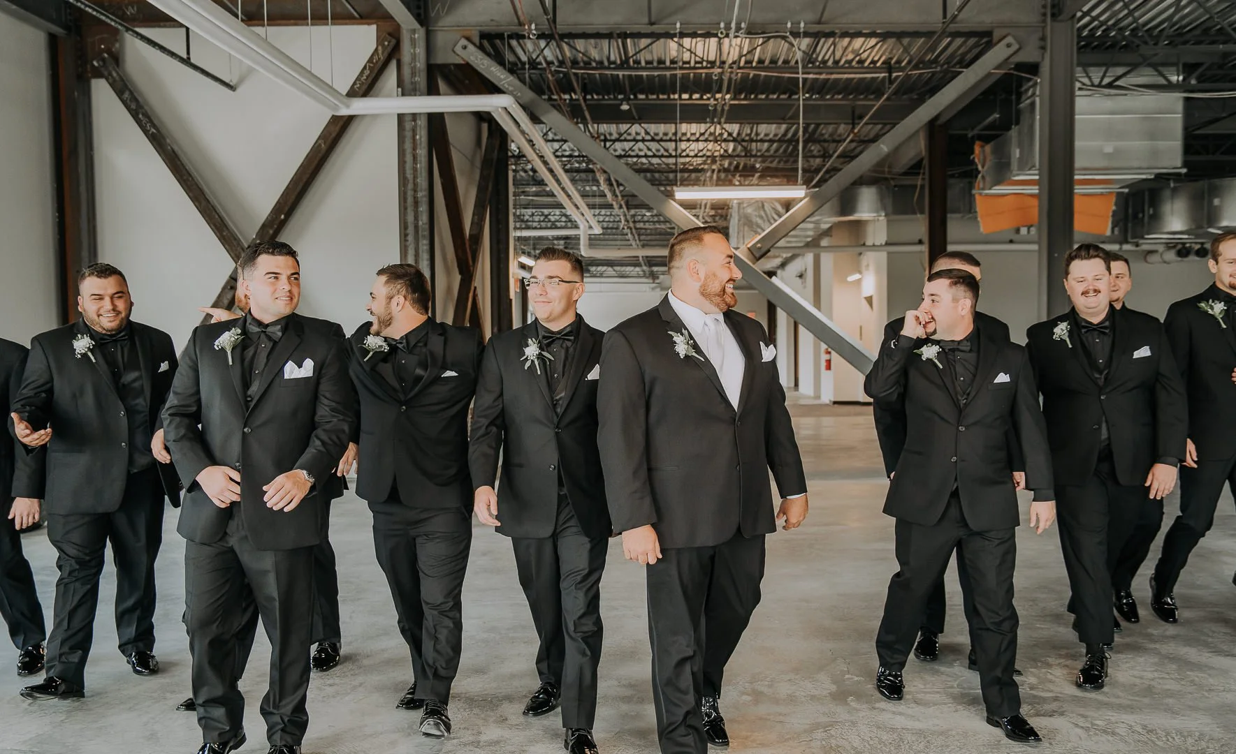 A group of men in black tuxedos walking together in an industrial-style indoor space, smiling and chatting.