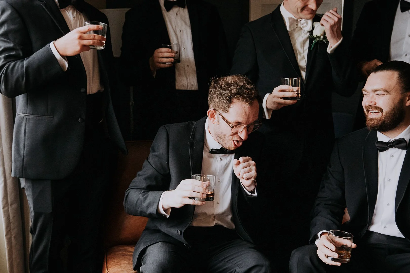 Group of men in tuxedos having drinks and laughing at a formal gathering.