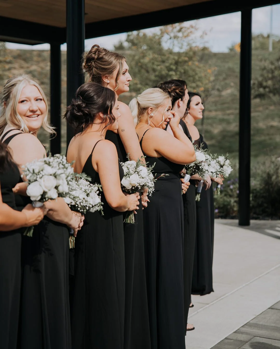 Group of women dressed in black bridesmaids' dresses holding bouquets at an outdoor wedding ceremony.