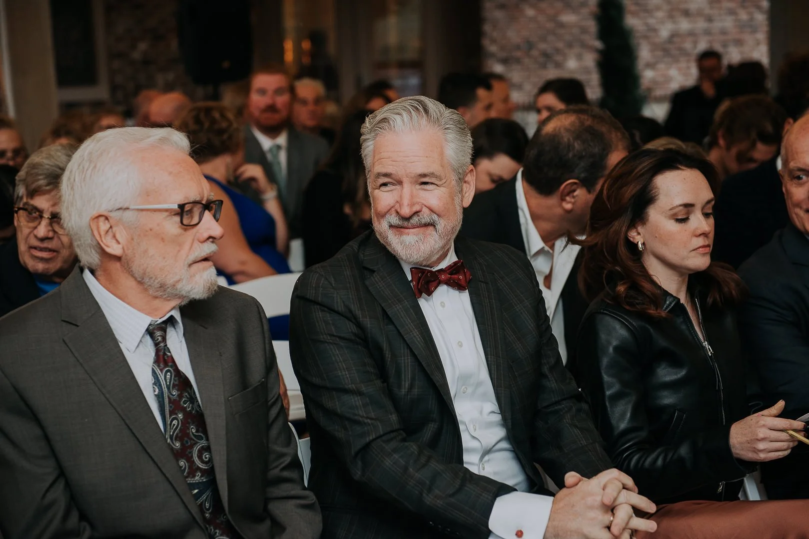 A group of formally dressed people attending an event, sitting in rows, in a room with brick walls. The focus is on a smiling older man with a beard and bow tie, sitting between a man with glasses and a woman in a black leather jacket.