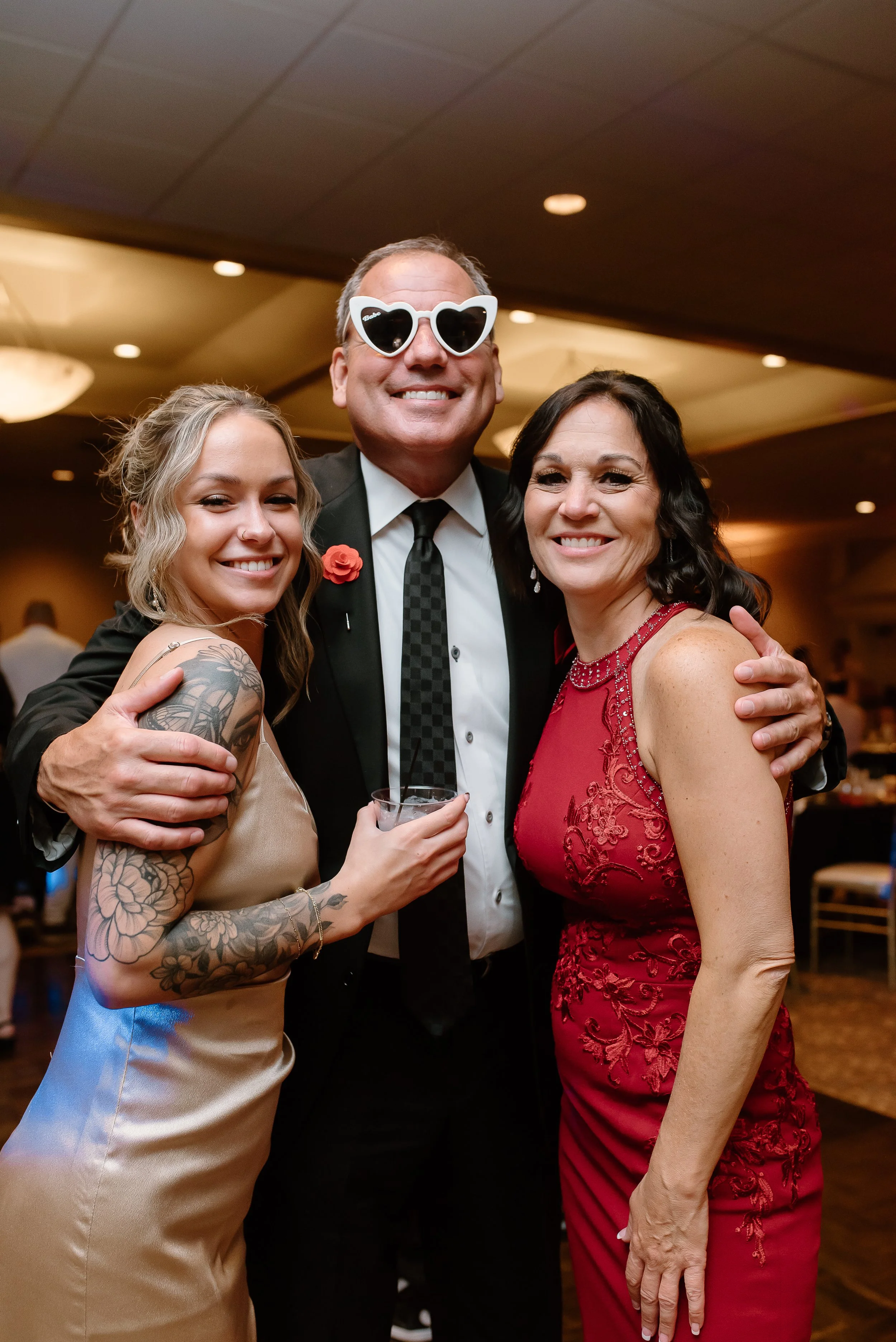 Three people smiling and posing for a photo at an indoor event, with one wearing white sunglasses and holding a drink, two women dressed elegantly, and a man in a suit.