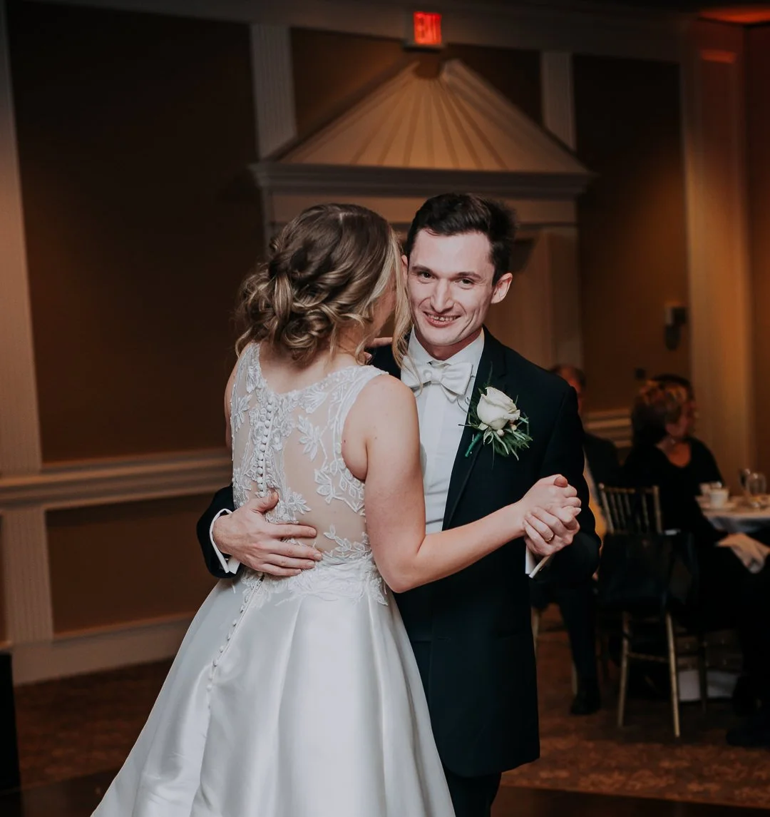 A bride and groom dancing at their wedding reception, smiling at each other in an elegant room with guests seated at tables in the background.