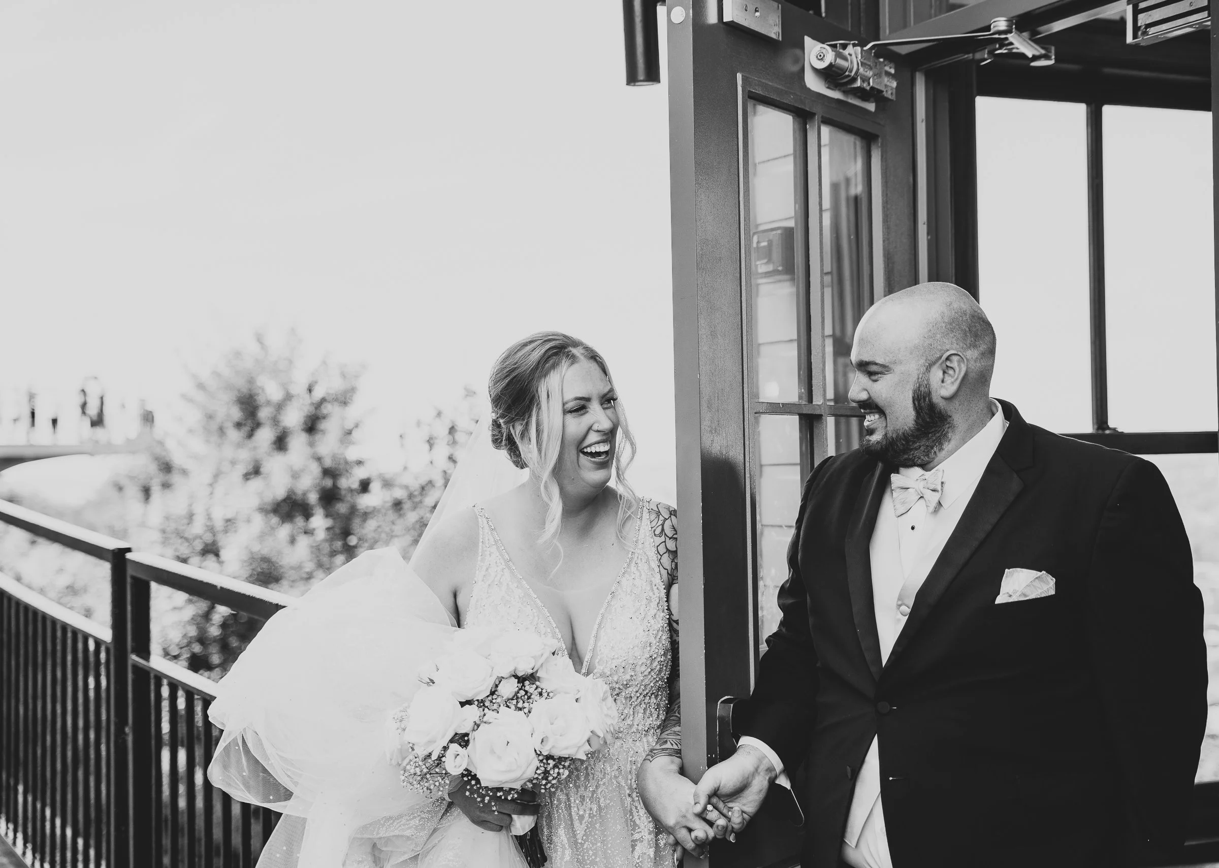 A bride and groom holding hands, smiling, standing outside near a door, with a woman in the background smiling and looking at them, in black and white.