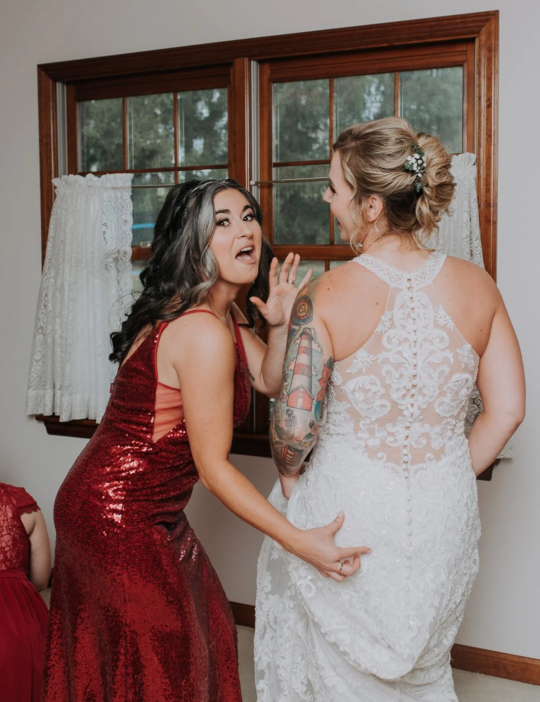 Two women in wedding attire sharing a moment inside a room with a wooden framed window behind them.