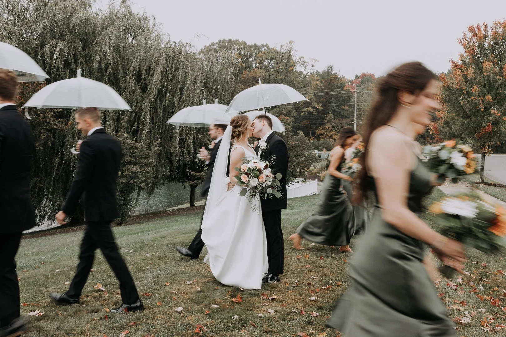 A bride and groom kiss under an umbrella at an outdoor wedding, surrounded by bridesmaids and groomsmen holding umbrellas, with autumn trees and a lake in the background.