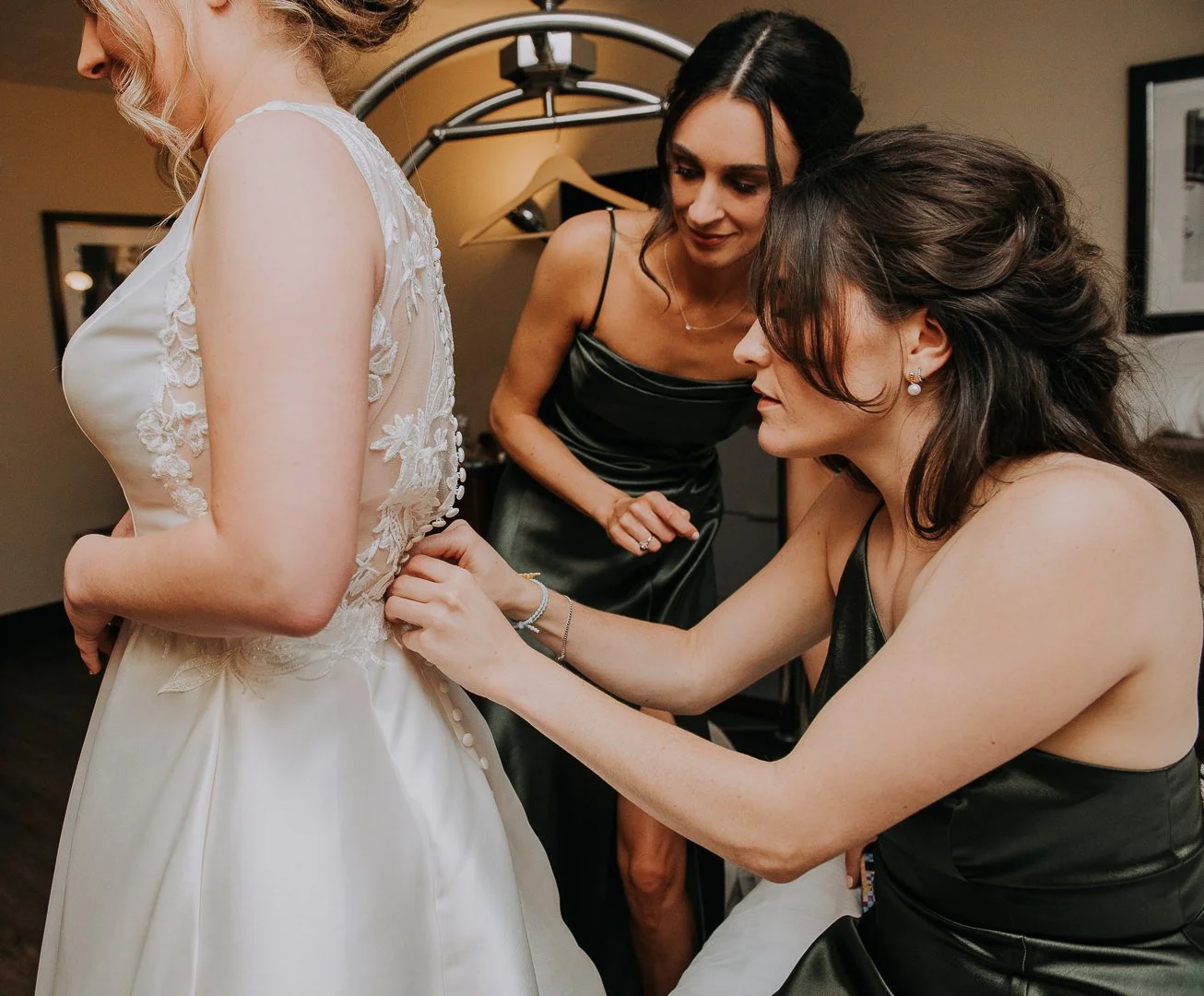 A bride in a white wedding dress is getting help with her dress from two women in black satin dresses, one kneeling and the other standing, inside a room.