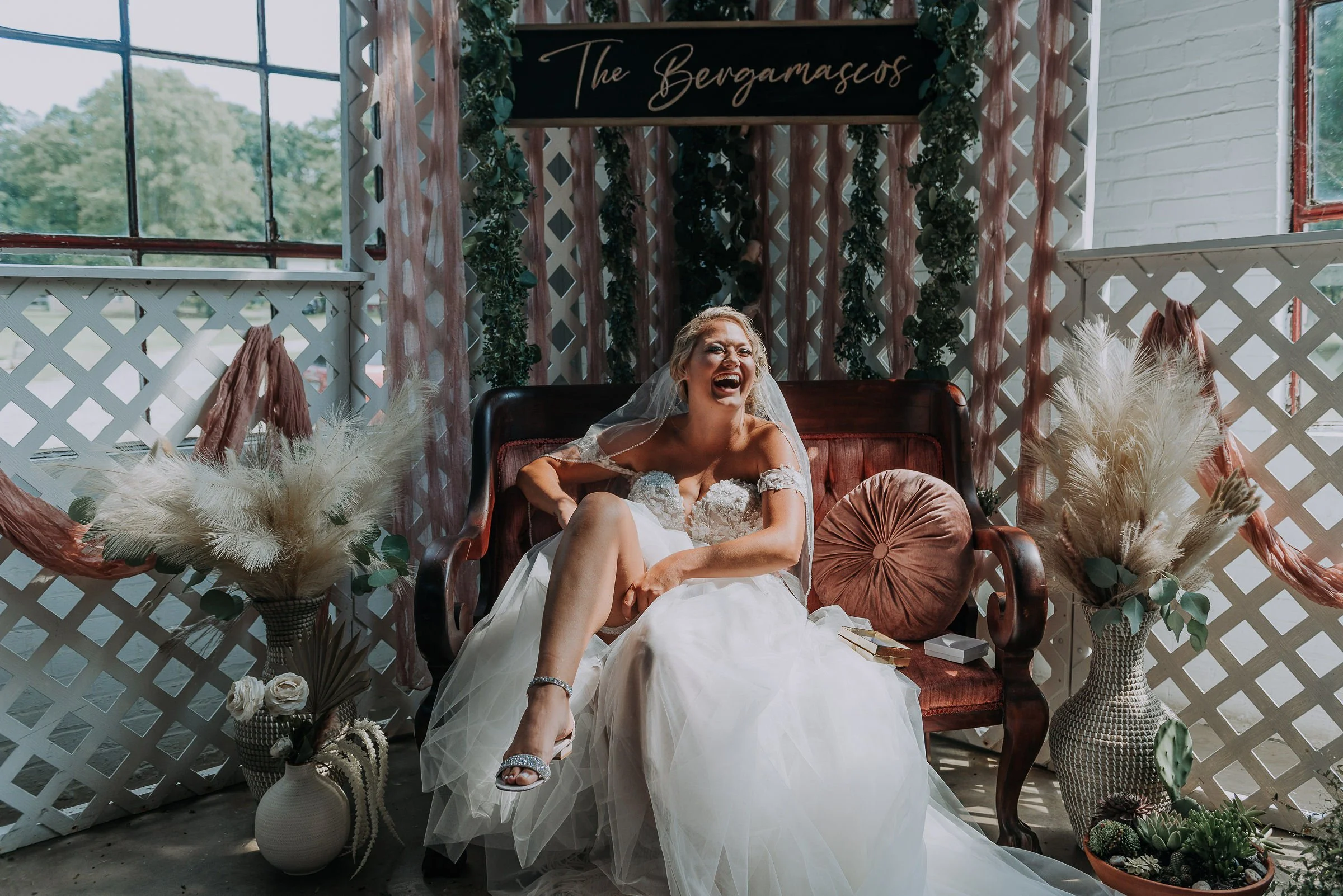 A bride in a wedding dress sitting on a wooden loveseat, laughing joyfully, with one leg raised. The setting is decorated with flower arrangements, pampas grass, vases, and drapes, with a sign reading 'The Bergamasco' hanging above.