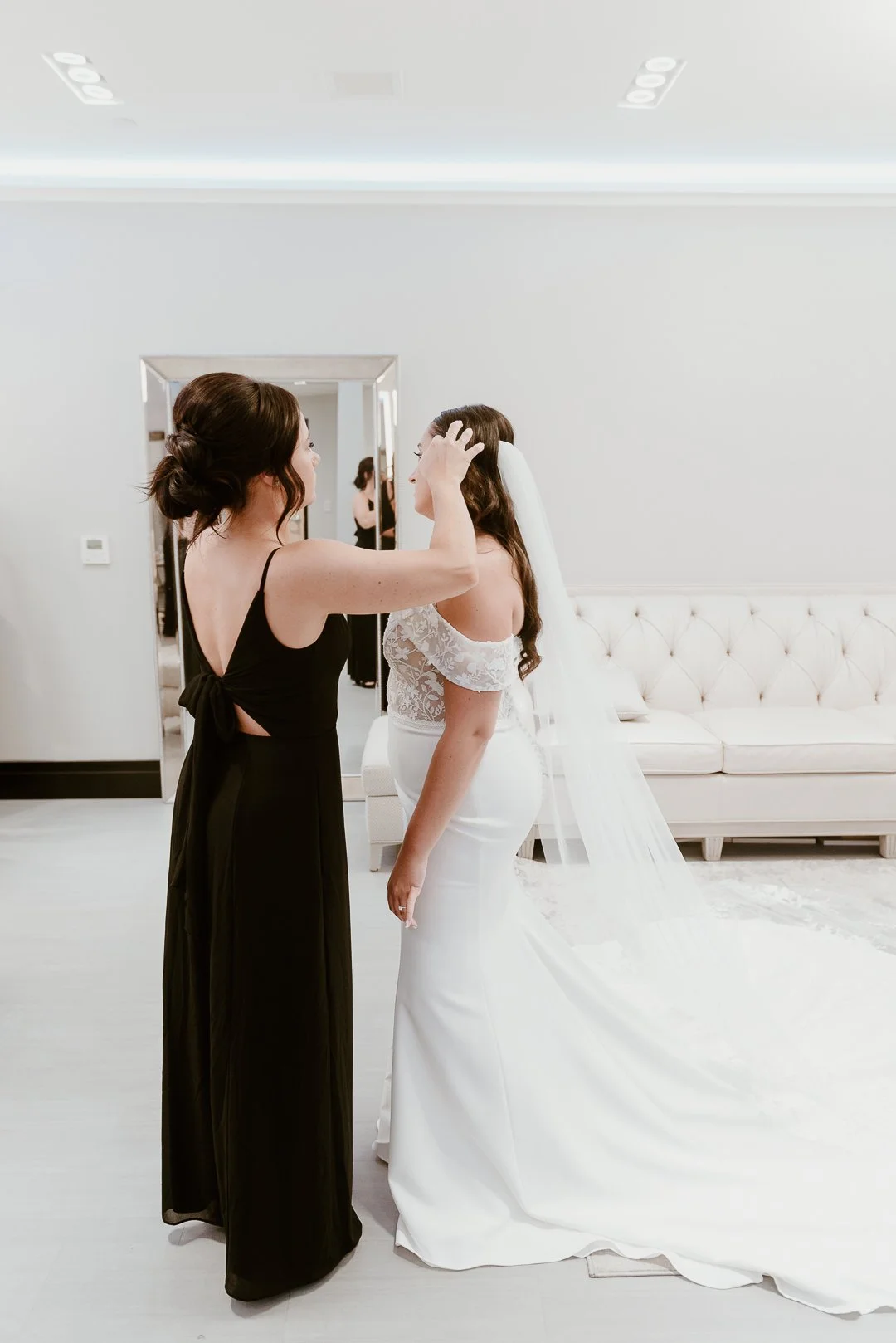 A bride in a white wedding dress with lace details is standing in front of a woman in a black dress, who is adjusting her hair in a modern, minimal room with a white tufted sofa and mirror.