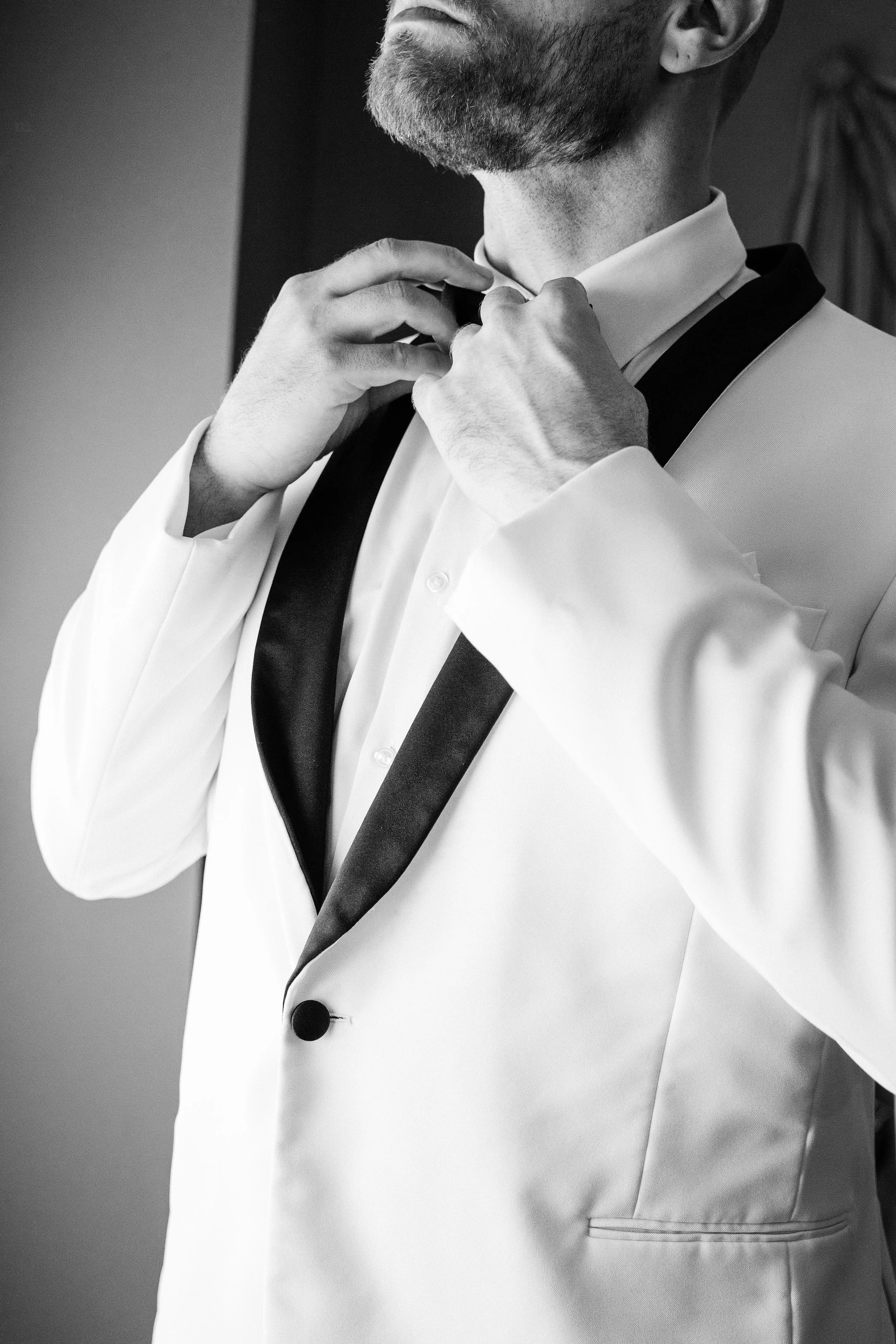 A man adjusting his bow tie in a tuxedo, viewed from the chest up, tight framing, black and white.