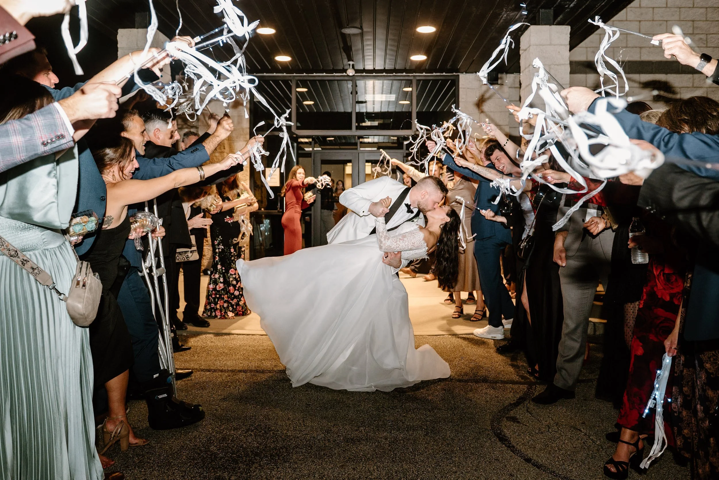 A newlywed couple sharing a kiss under a shower of white streamers and confetti with wedding guests forming a corridor around them.