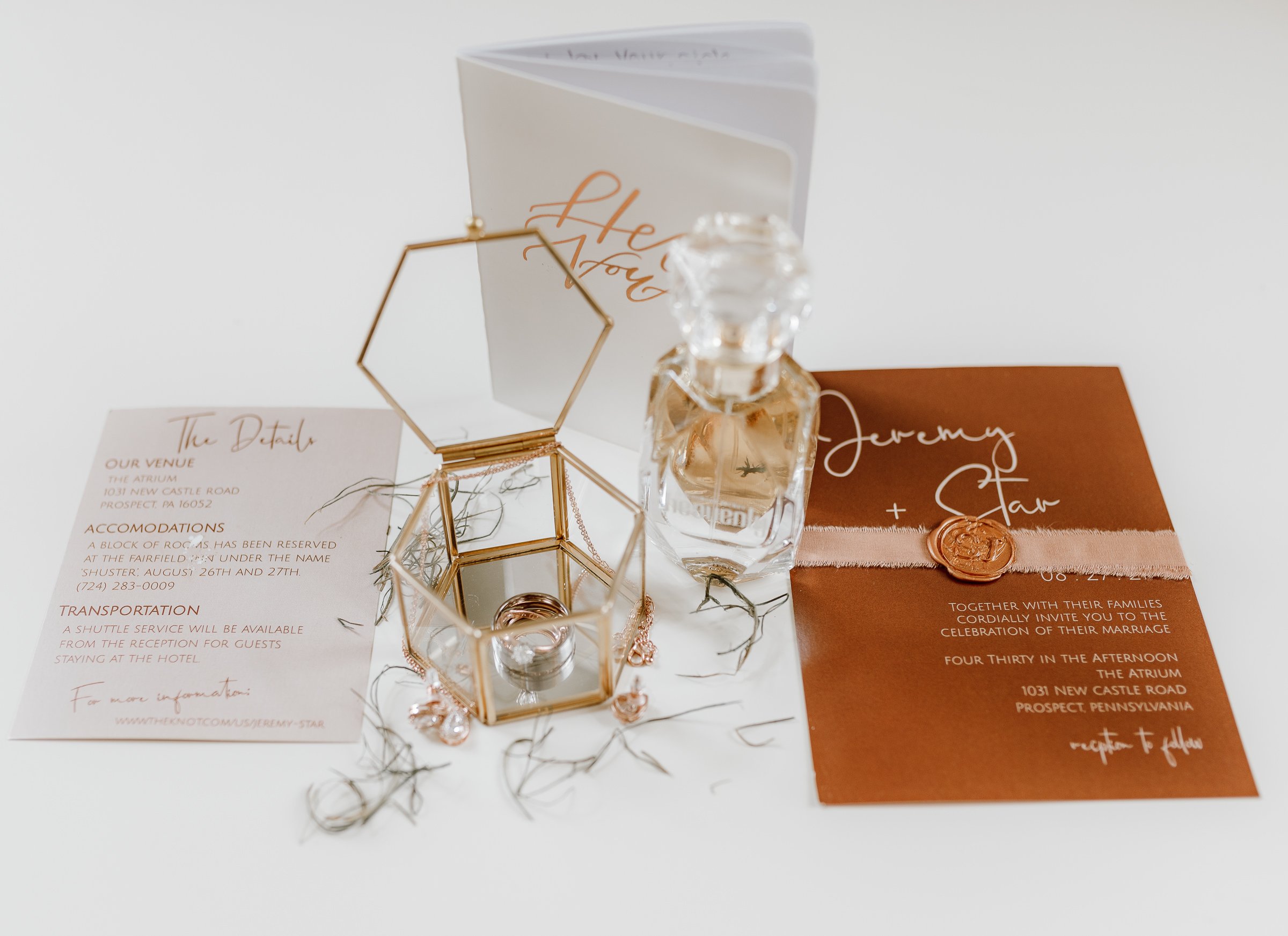 Wedding invitation, glass perfume bottle, decorative gold geometric mirror, and wedding details card on a white surface.