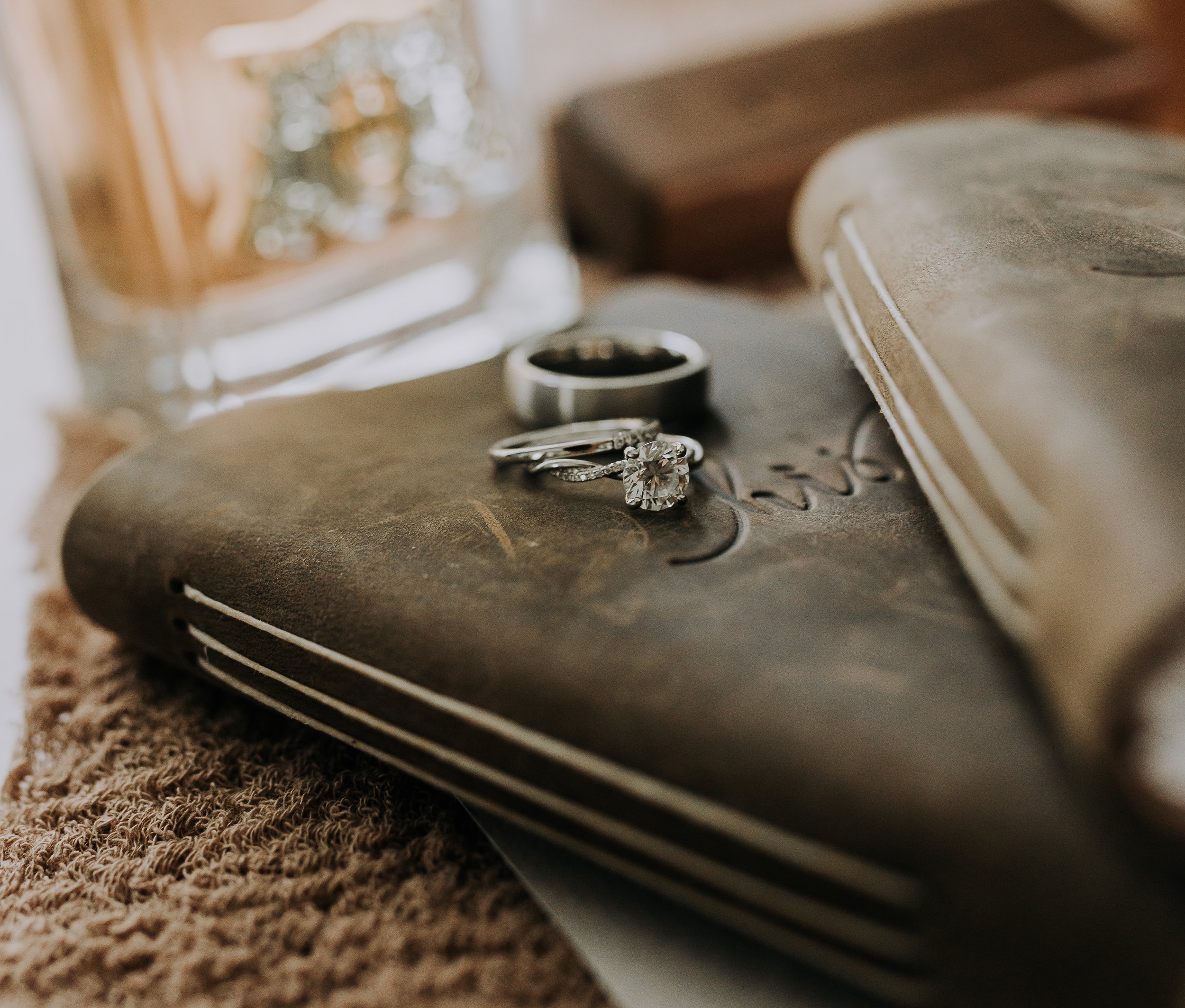 Close-up of three rings, including an engagement ring with a large diamond, on a textured surface near a book and a blurred glass container in the background.