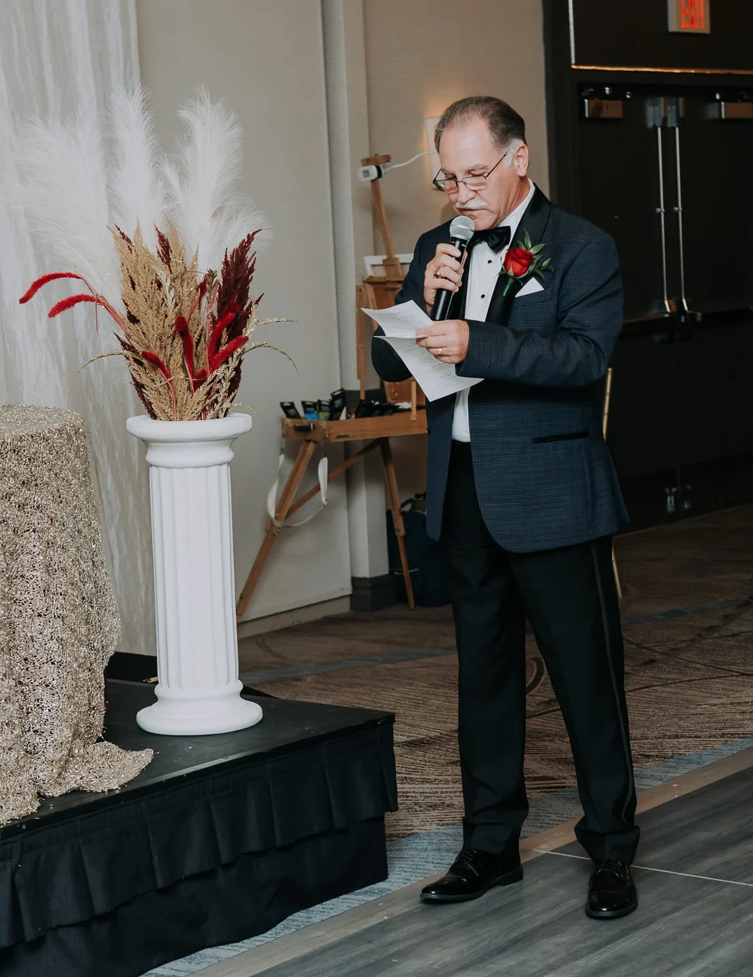 A man in a tuxedo with a red rose boutonniere reads from a paper while holding a microphone at an indoor event.