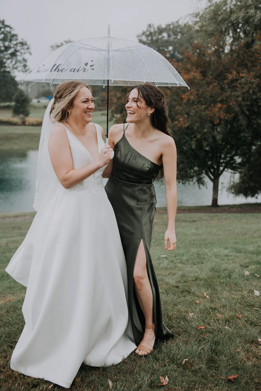 Two women standing outdoors under a clear umbrella, showing a joyful moment on a rainy day. One is wearing a white wedding dress with a veil, and the other is in a dark green satin dress with a high slit. They are smiling and laughing together near a