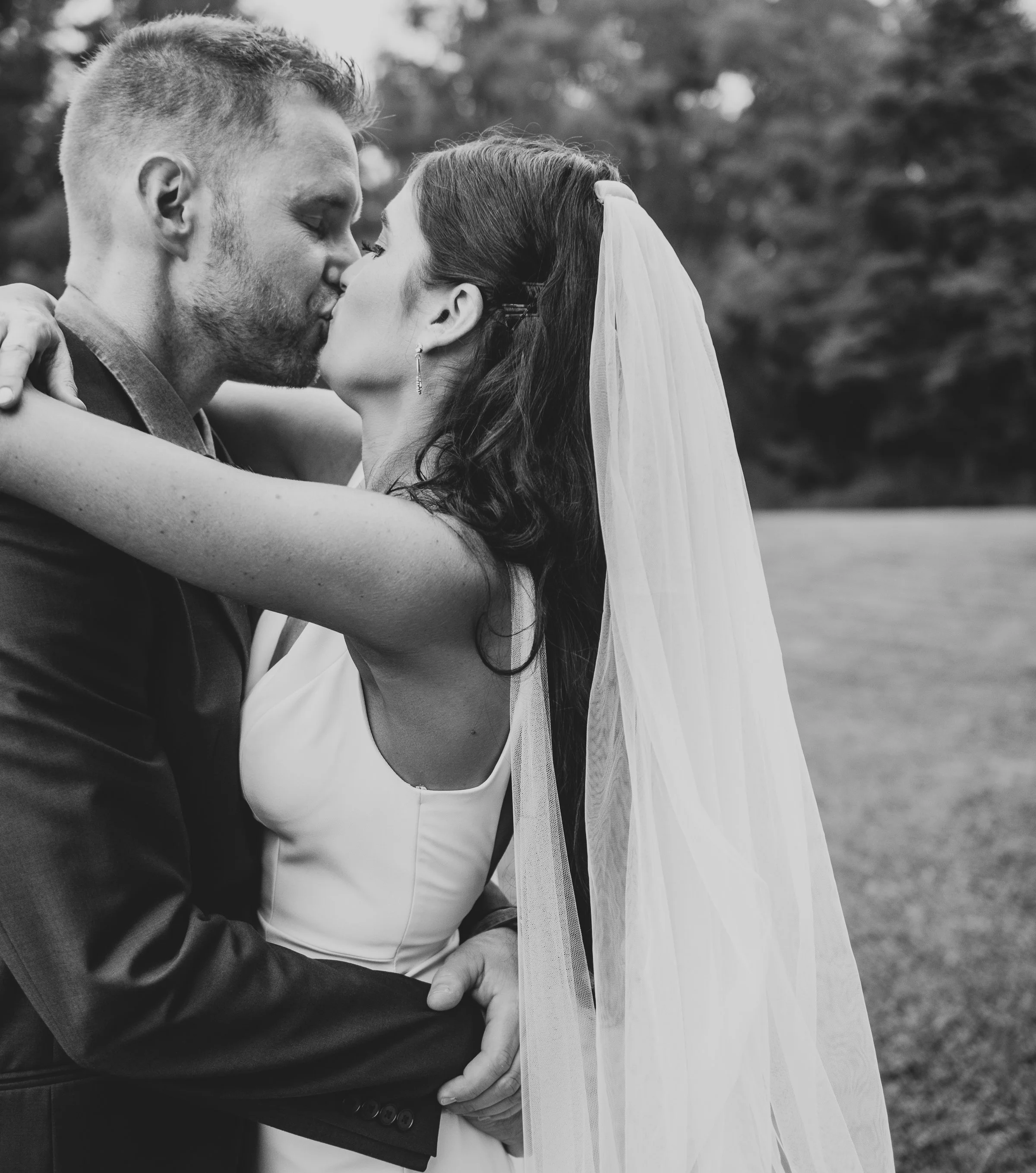 A black and white photo of a bride and groom kissing, embracing each other closely outdoors. The groom has short hair and a beard, wearing a tuxedo. The bride has long hair, wears a veil, and a white dress. The background is blurred with trees.