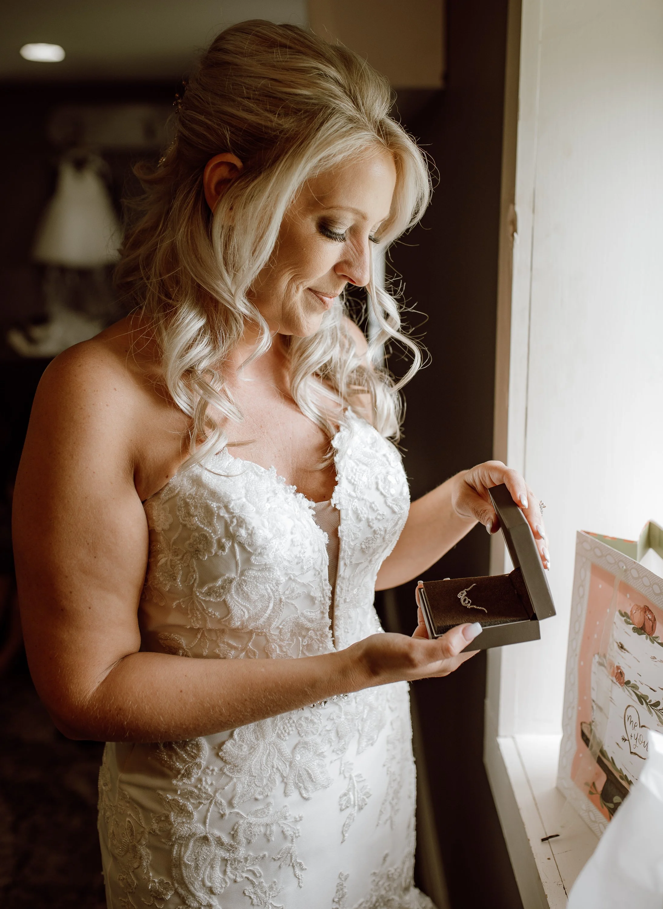 A woman in a wedding dress looking at a jewelry box with a necklace inside, standing near a window.