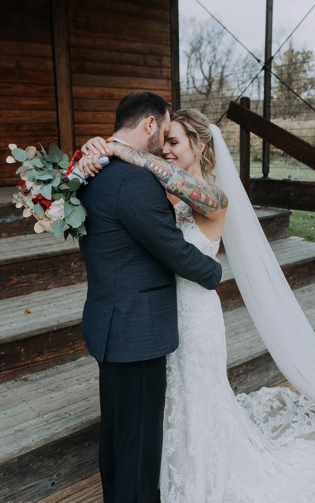 A newlywed couple embraces outside a wooden building, smiling and happy, with the bride wearing a white lace wedding gown and veil, and the groom in a dark suit. The bride has tattoos on her arms and is holding a bouquet of eucalyptus leaves and red 
