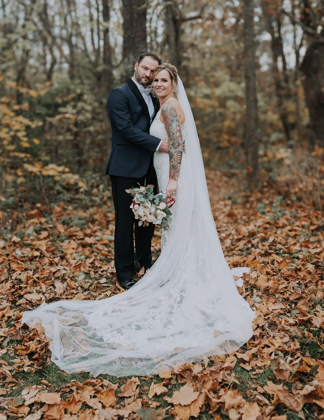 A bride and groom standing in a forest with autumn leaves, dressed in wedding attire. The bride has tattoos on her arms and holds a bouquet, and the groom wears a dark suit. They are smiling and embracing each other.