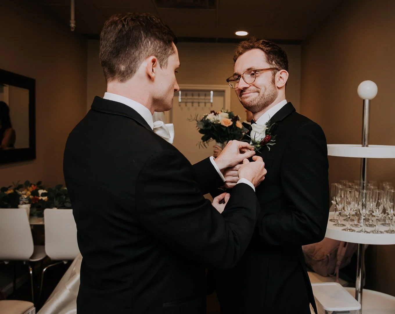 Two men in tuxedos participating in a wedding ceremony, with one helping the other with a boutonniere, in an indoor setting with glasses on a table and a bouquet in the background.