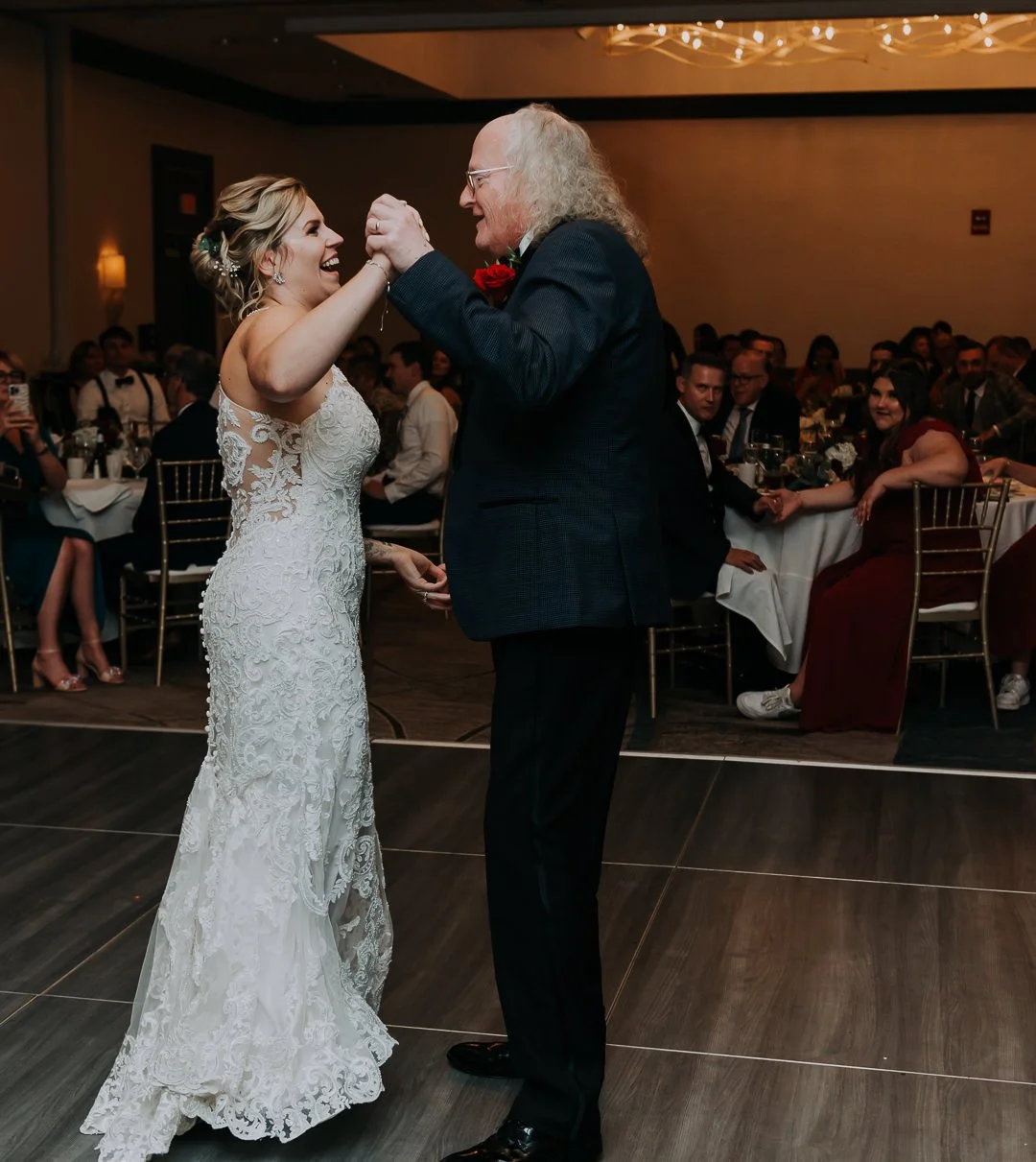A bride and an older man, possibly her father, share a dance at a wedding reception. The bride is wearing a white lace wedding dress and has blonde hair, while the man has long gray curly hair, glasses, and is dressed in a dark suit with a red bouton