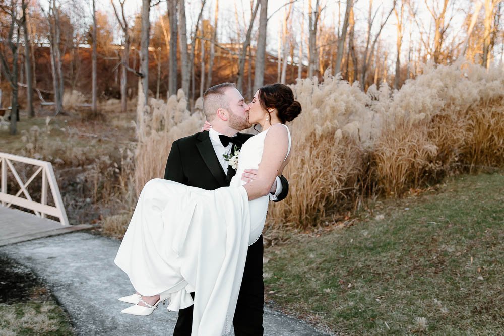 A bride in a white wedding dress being carried by a groom in a black tuxedo, both sharing a kiss outdoors in a scenic autumn setting.