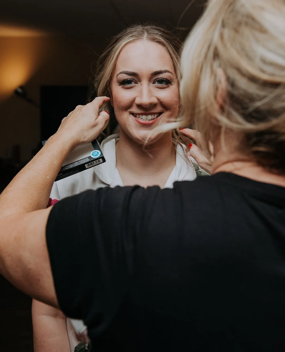 A woman with blonde hair getting her makeup done by another woman with blonde hair, smiling and looking pleased.