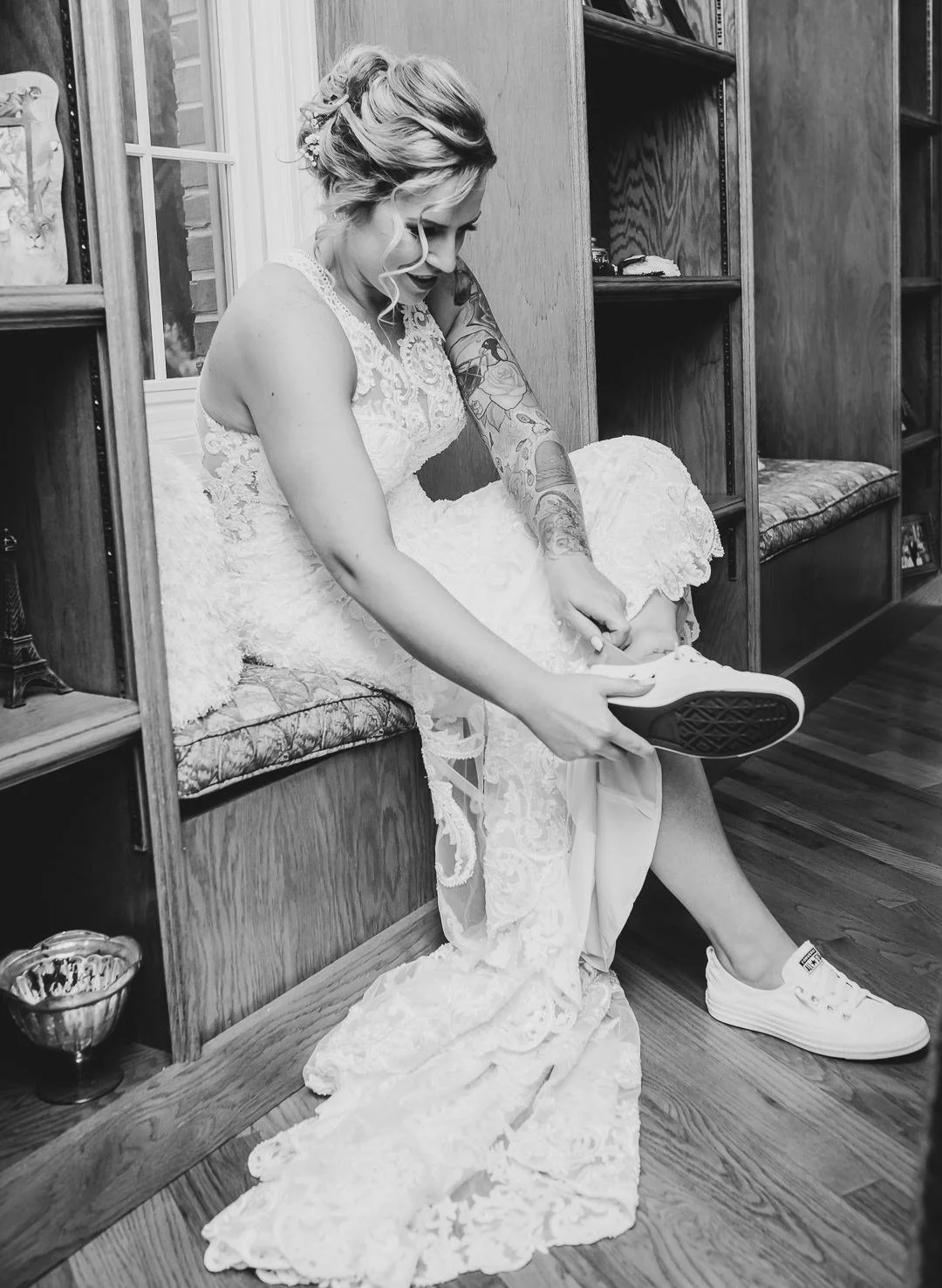 A bride in a lace wedding dress putting on sneakers while sitting on a built-in wooden bench in a room with wooden shelves and flooring.