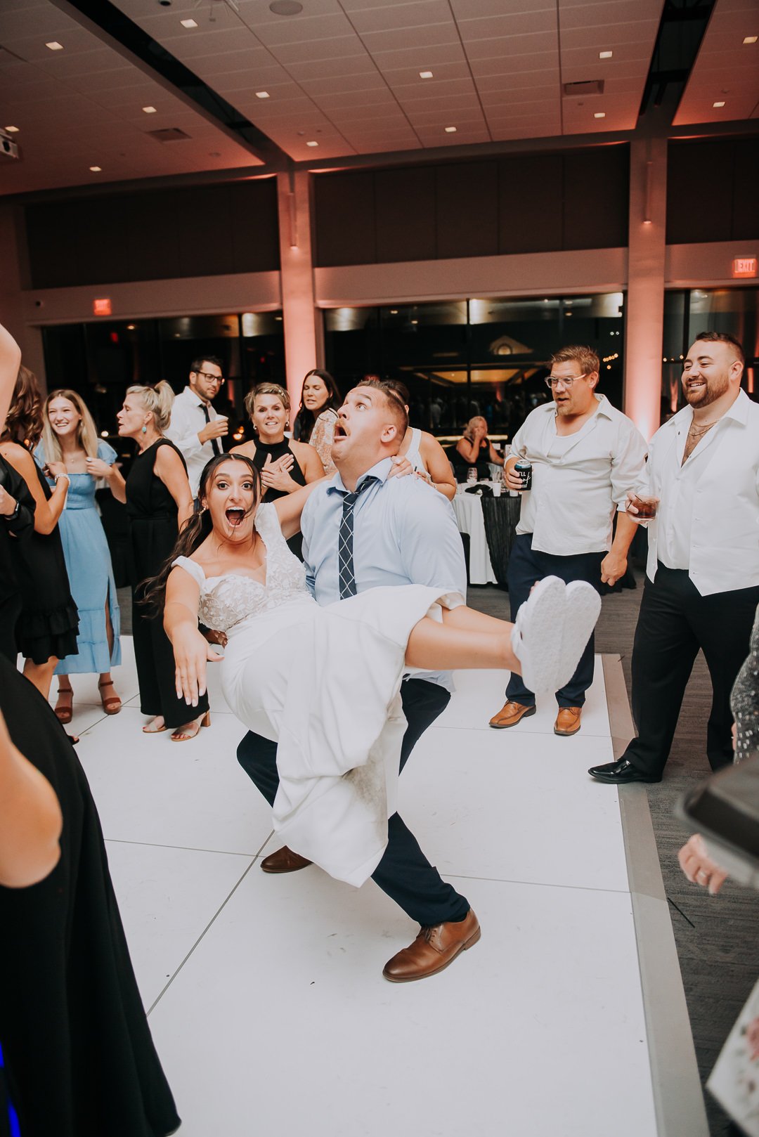 A groom carrying a joyful bride as they dance at a wedding reception with guests in the background.