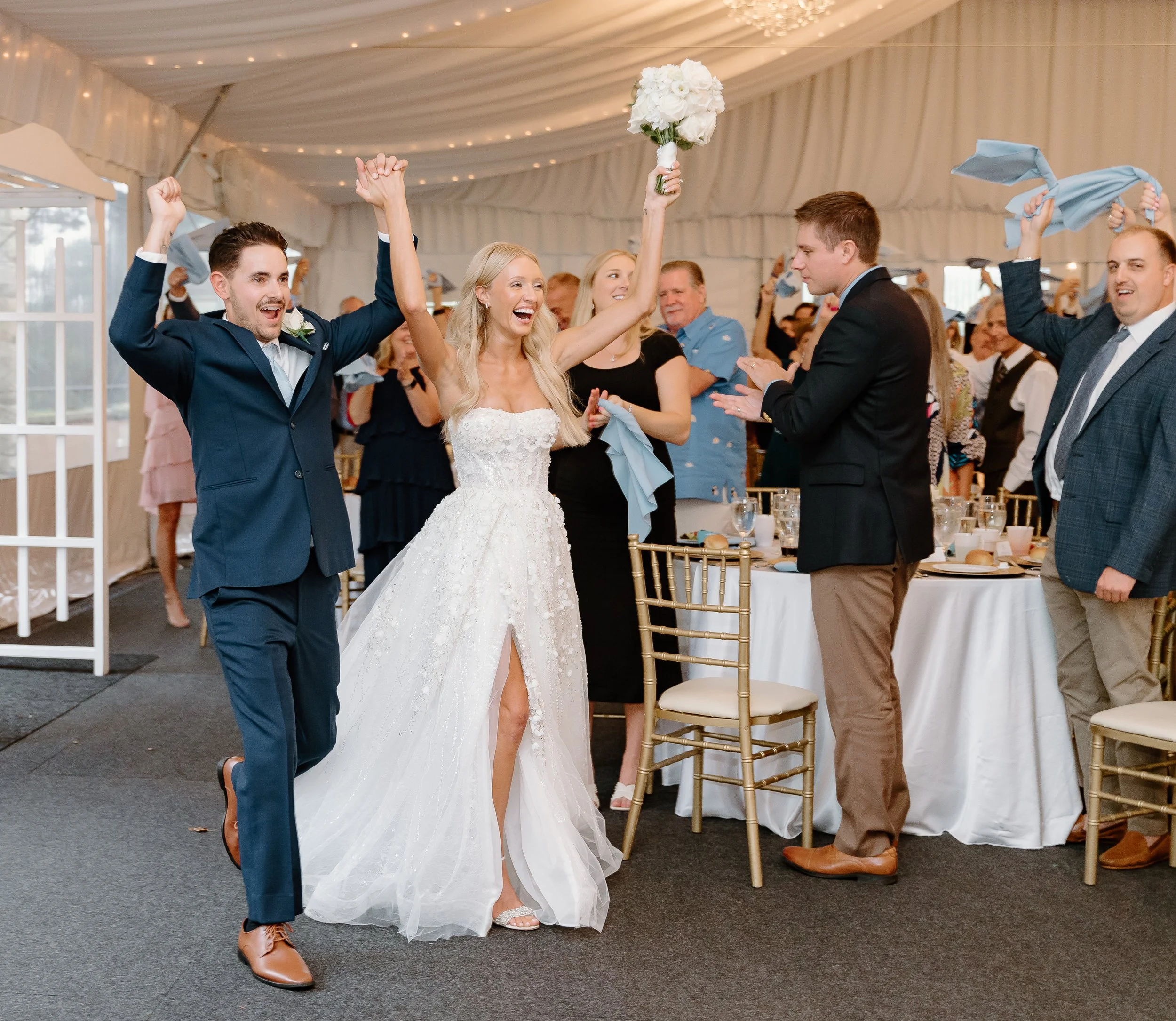 A wedding reception scene with a bride, groom, and guests celebrating indoors under a tent with string lights. The bride is holding a bouquet and smiling, while the groom is excited with raised arms.