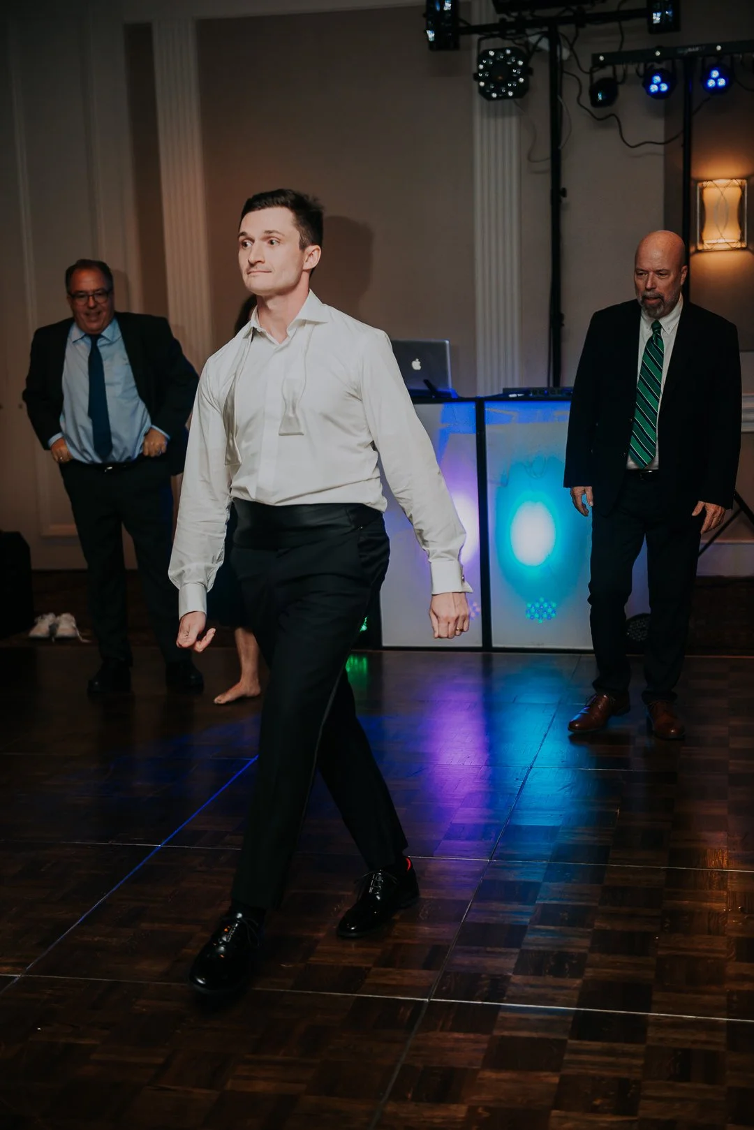 Man in white shirt and black pants dancing on wooden floor in a party setting with two men in suits in the background and a DJ setup with colorful lights.