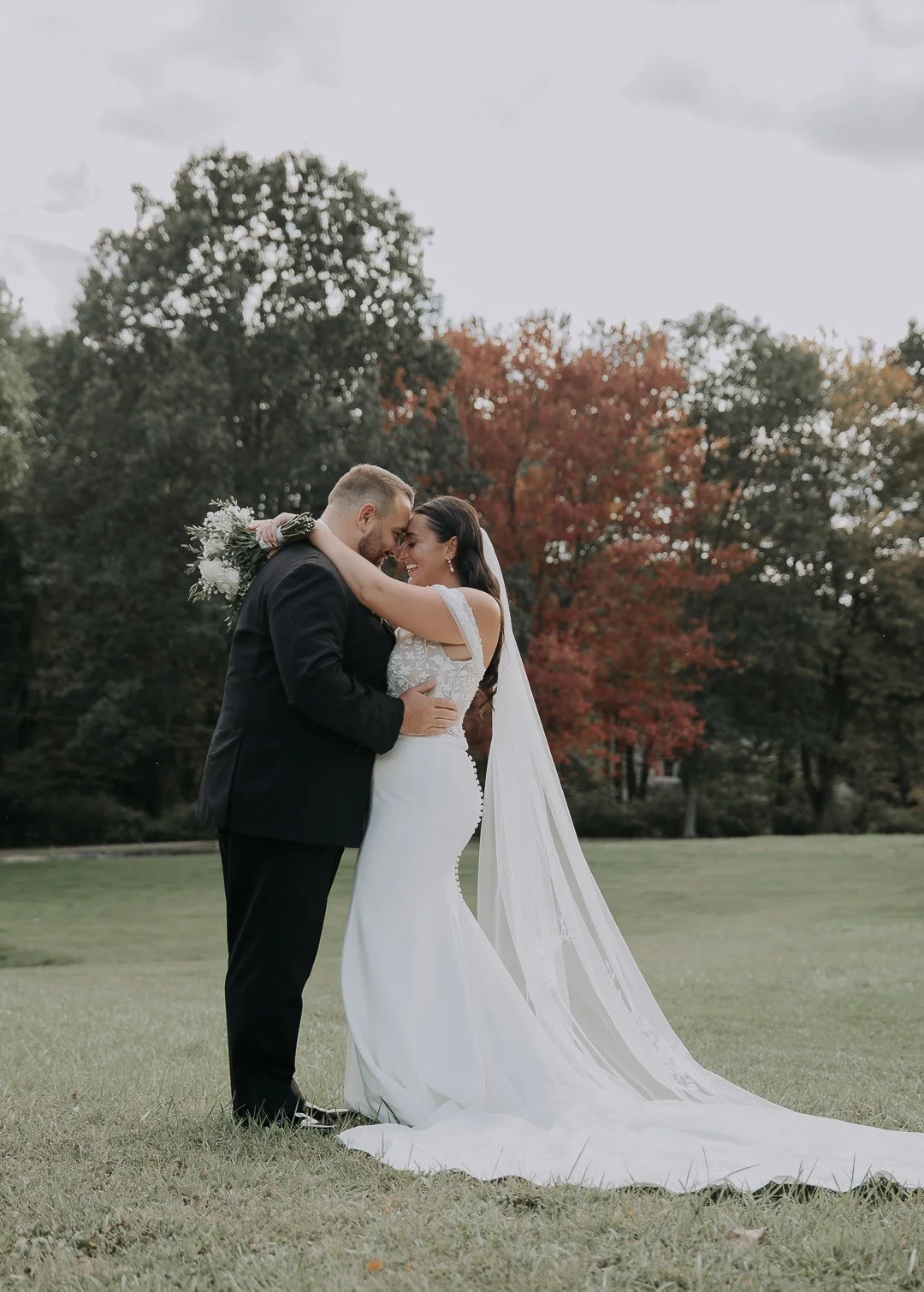 A bride and groom embracing outdoors on a grassy field with autumn trees in the background.