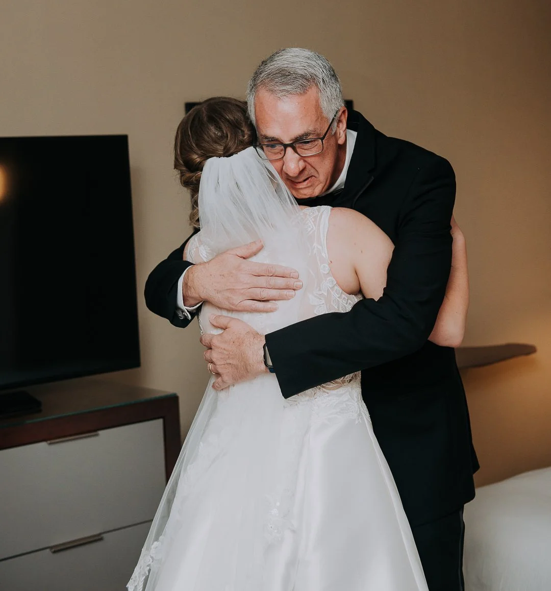 A bride hugging an older man, likely her father, in a hotel room, both emotional on her wedding day.