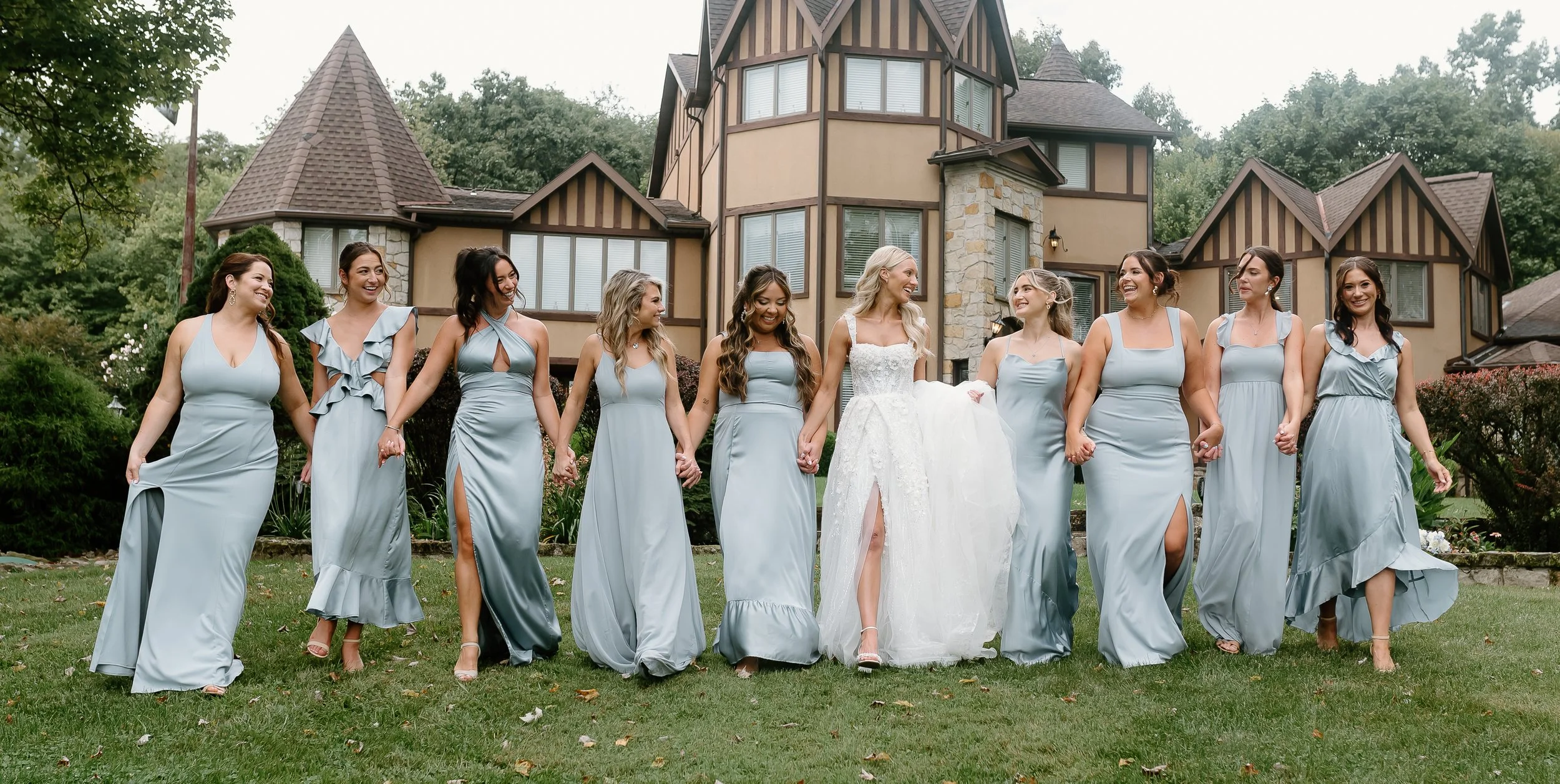 A group of eleven women, including a bride in a white wedding dress, holding hands and walking on a lawn in front of a large house with a stone and wood exterior. The women are dressed in matching light blue dresses and appear to be celebrating a wed