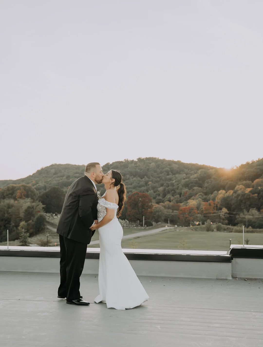 A newlywed couple sharing a kiss on a rooftop at sunset with a scenic view of trees and hills in the background.