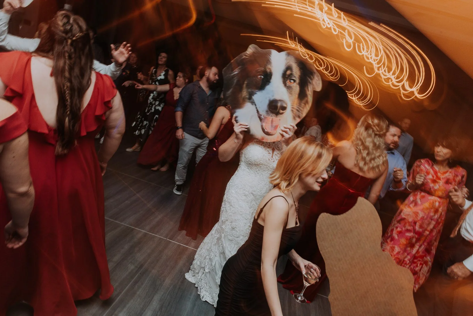 Party scene with people dancing, a woman holding a large dog face cutout, and vibrant light streaks overhead.