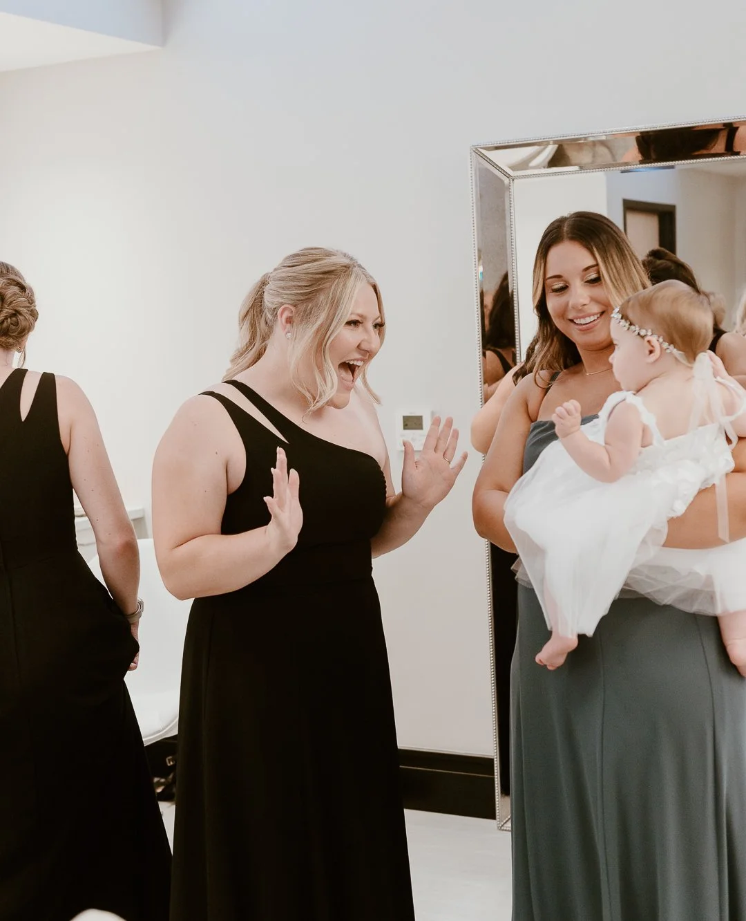Two women in black dresses are smiling and having fun while looking at a baby girl in a white dress being held by another woman in a gray dress. They are in a room with a large mirror.